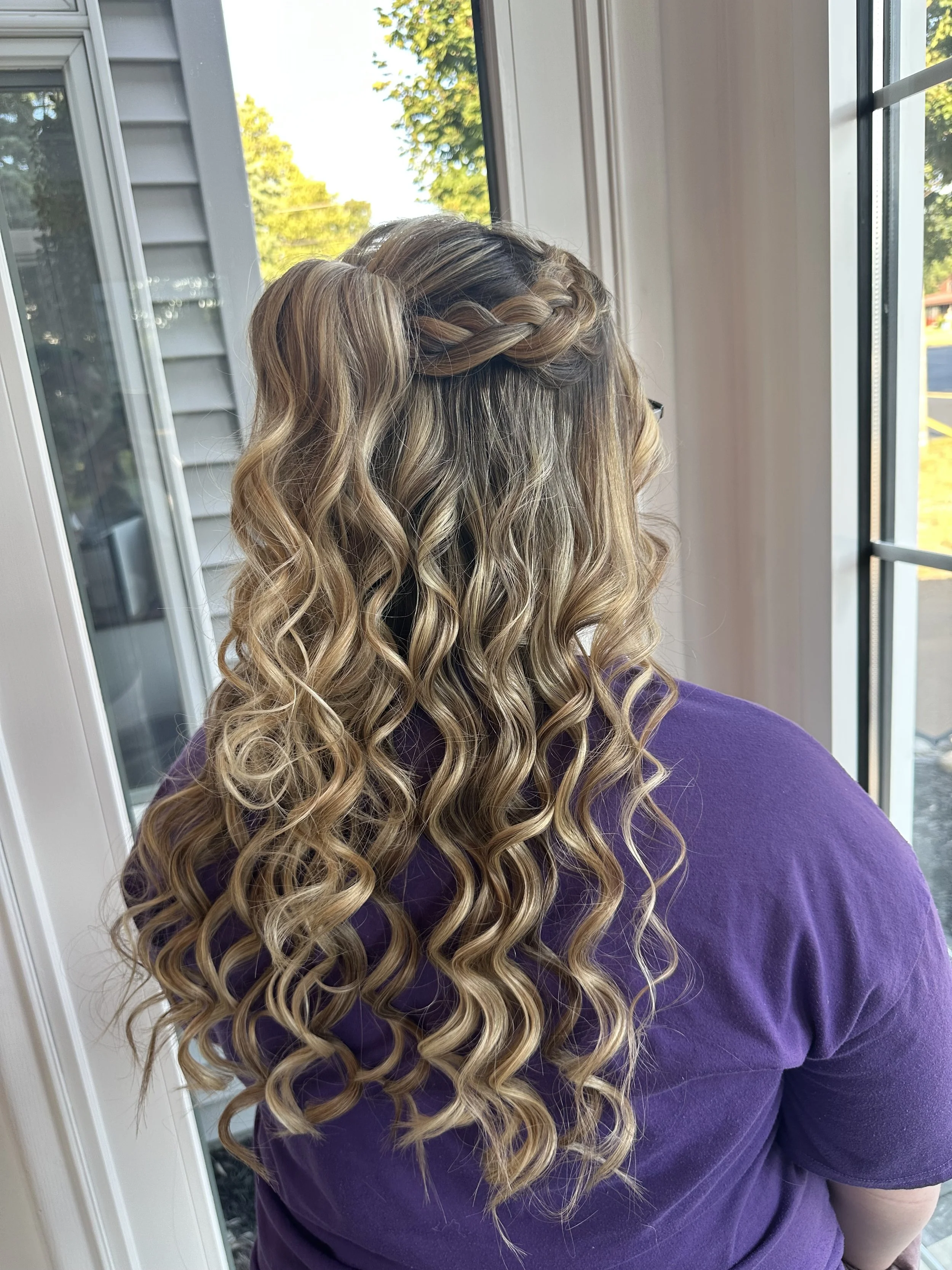 A woman with long, curly blonde hair styled with a braided crown, wearing a purple shirt, standing indoors near a window with green trees visible outside.