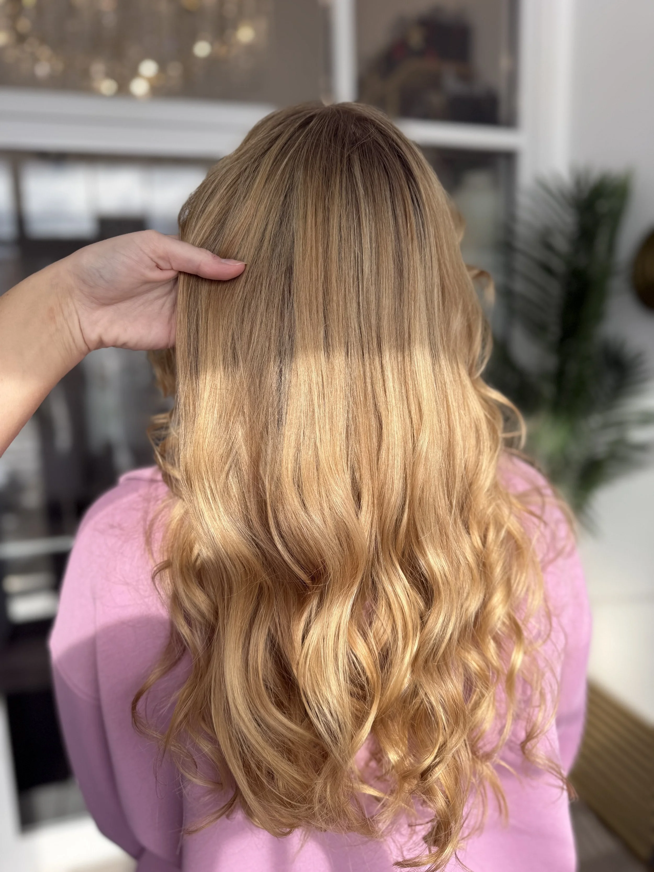 A person with long, wavy blonde hair wearing a pink shirt, with a hand gently touching the hair.