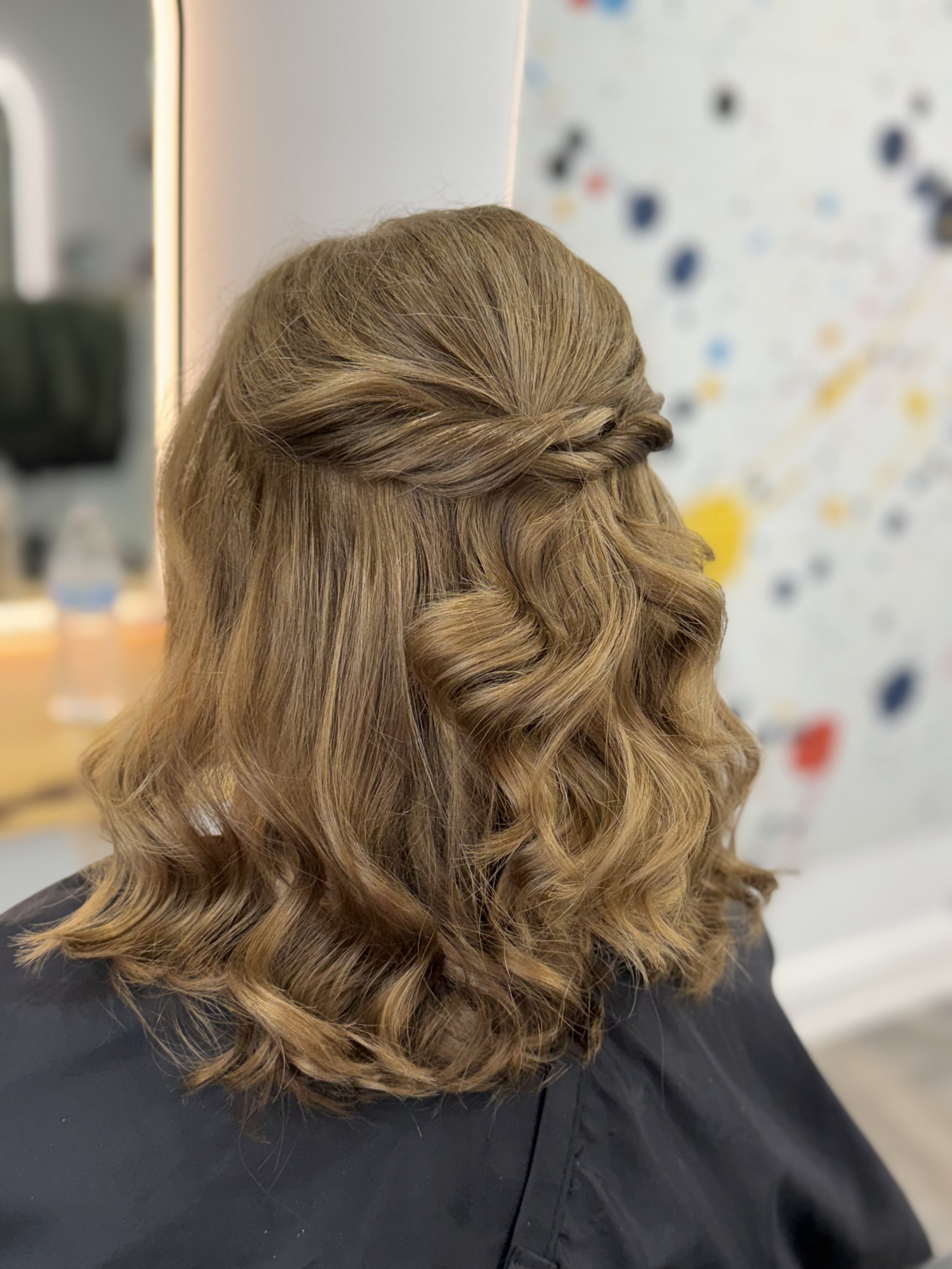 A person's head with styled golden or light brown hair featuring loose curls and a twisted strand across the front. The background is blurred with colorful dots and a white surface.