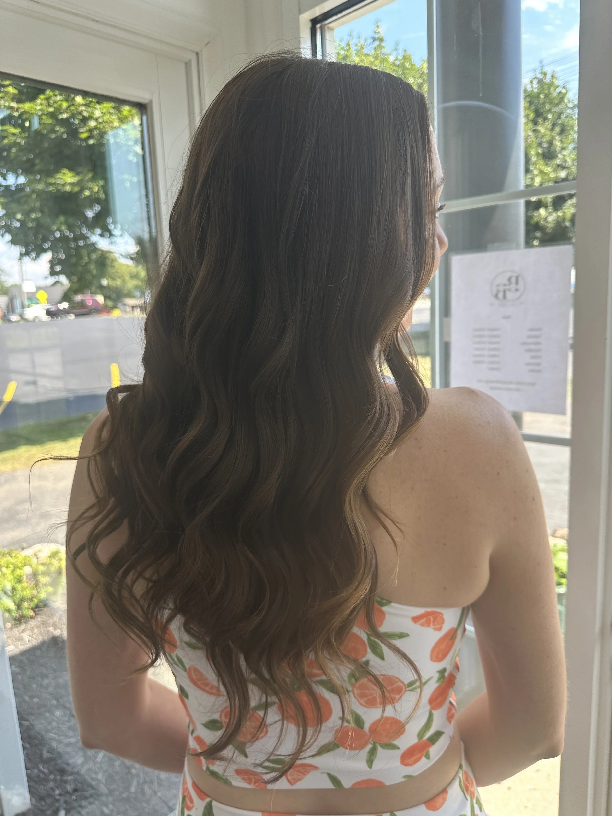 Woman with long brown curly hair standing near a door with glass panels, looking outside at a sunny day with green trees and parked cars.