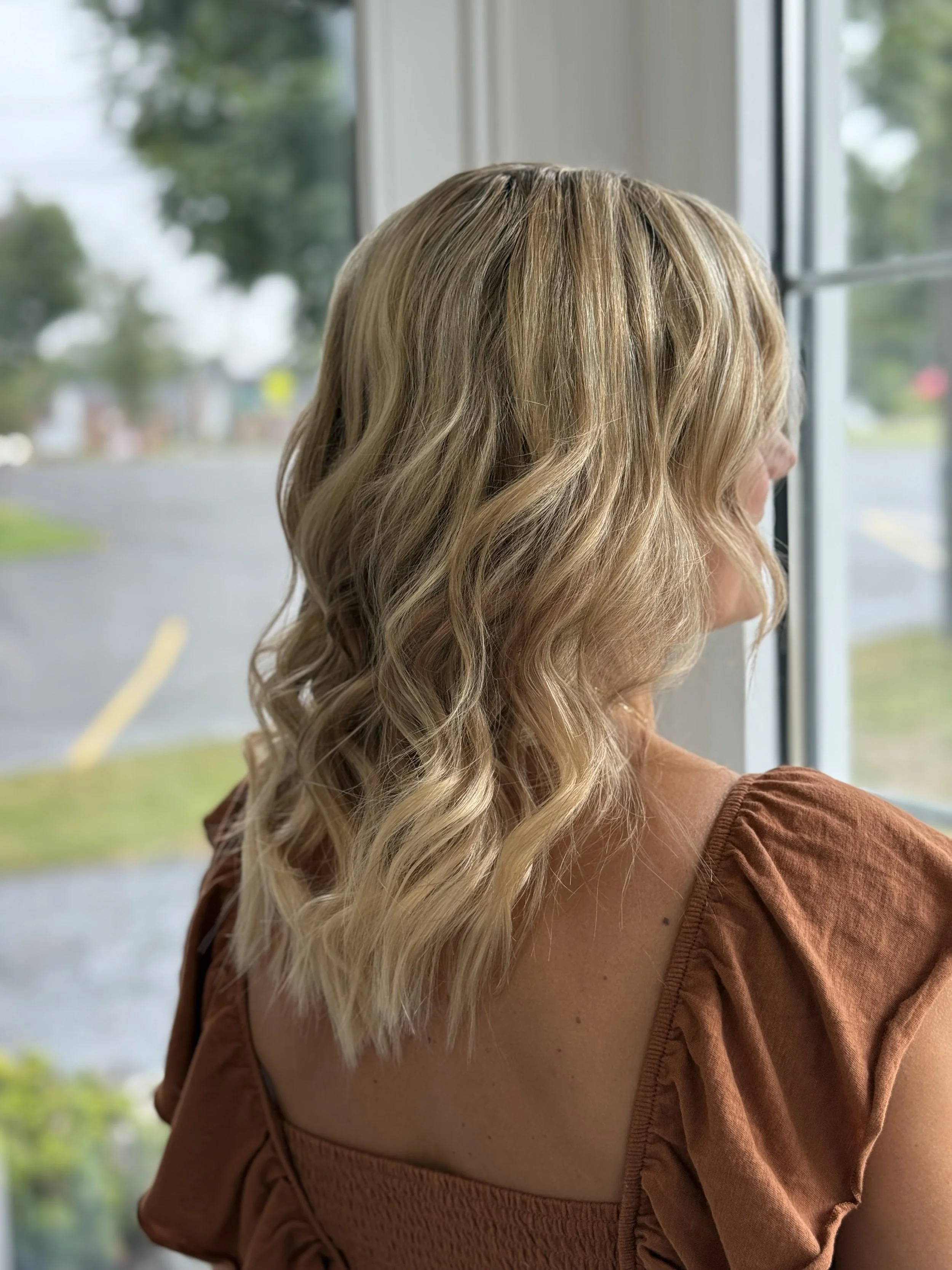 A woman with long, wavy blonde hair looking out a window, wearing a brown top with puffed sleeves.