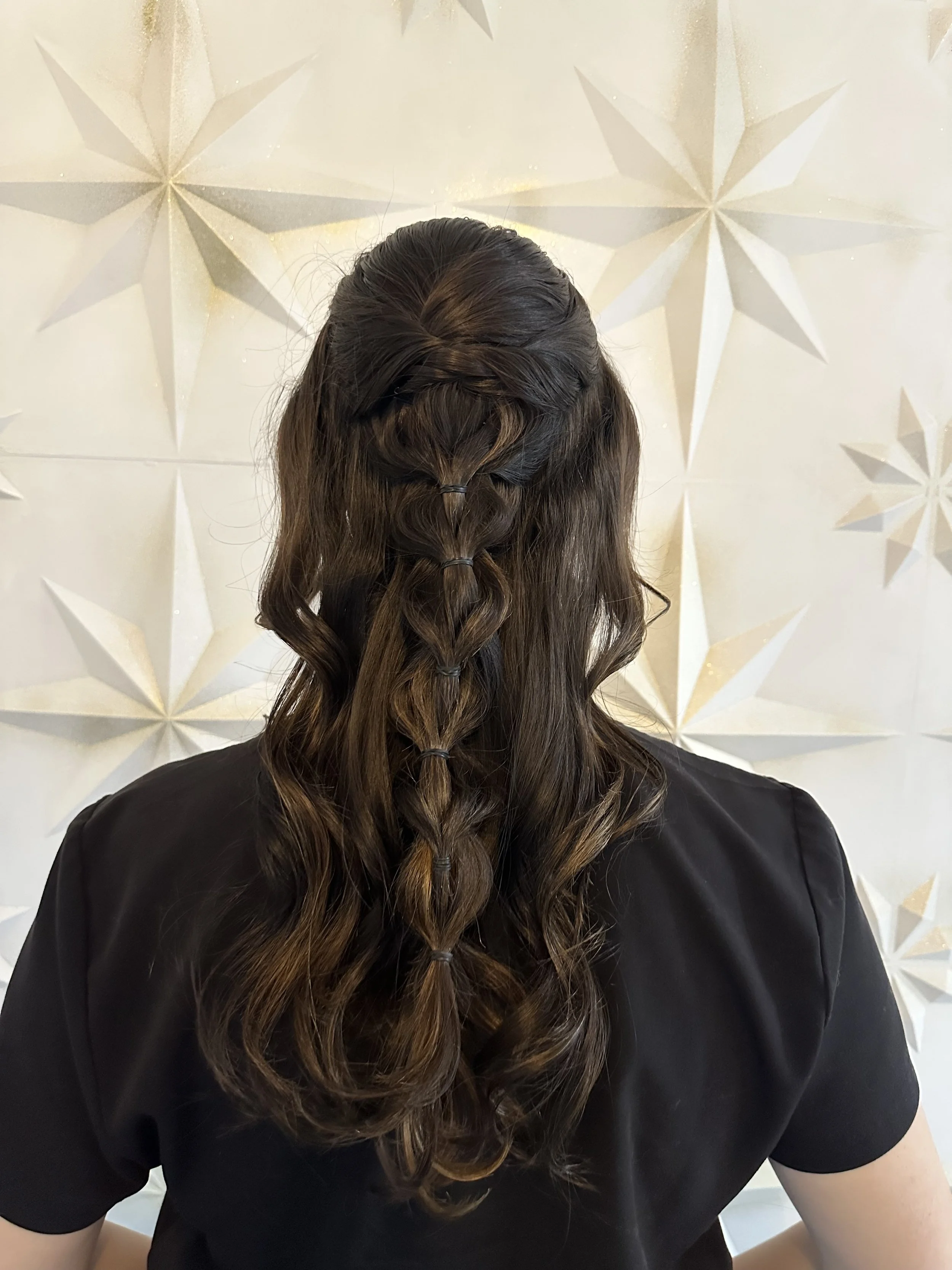 A person with long, dark brown, curled hair styled in a waterfall braid, wearing a black top, standing in front of a white wall decorated with large gold and white star-shaped ornaments.