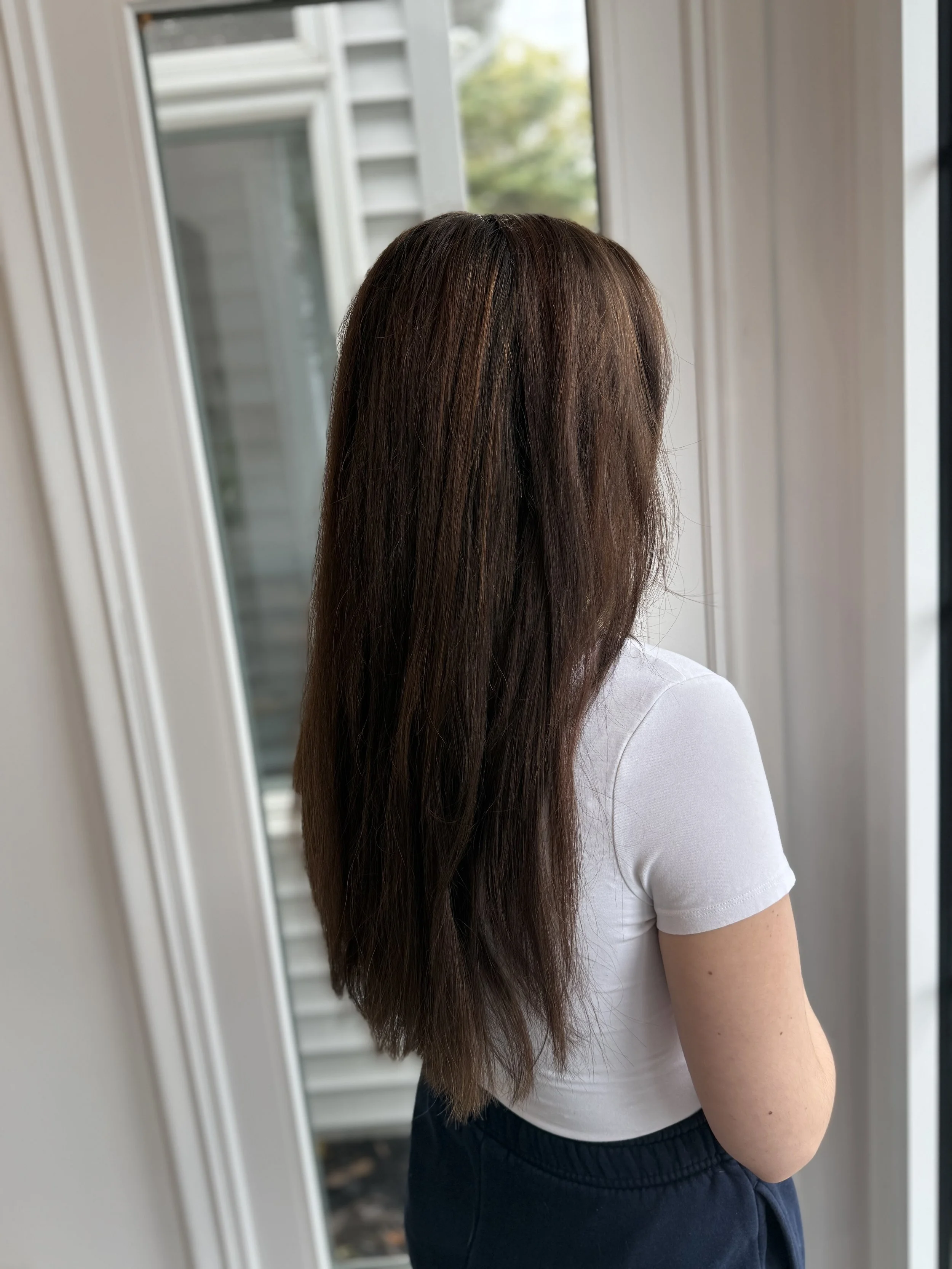 A woman with long, straight brown hair standing inside near a window, wearing a white shirt and dark pants, looking outside.