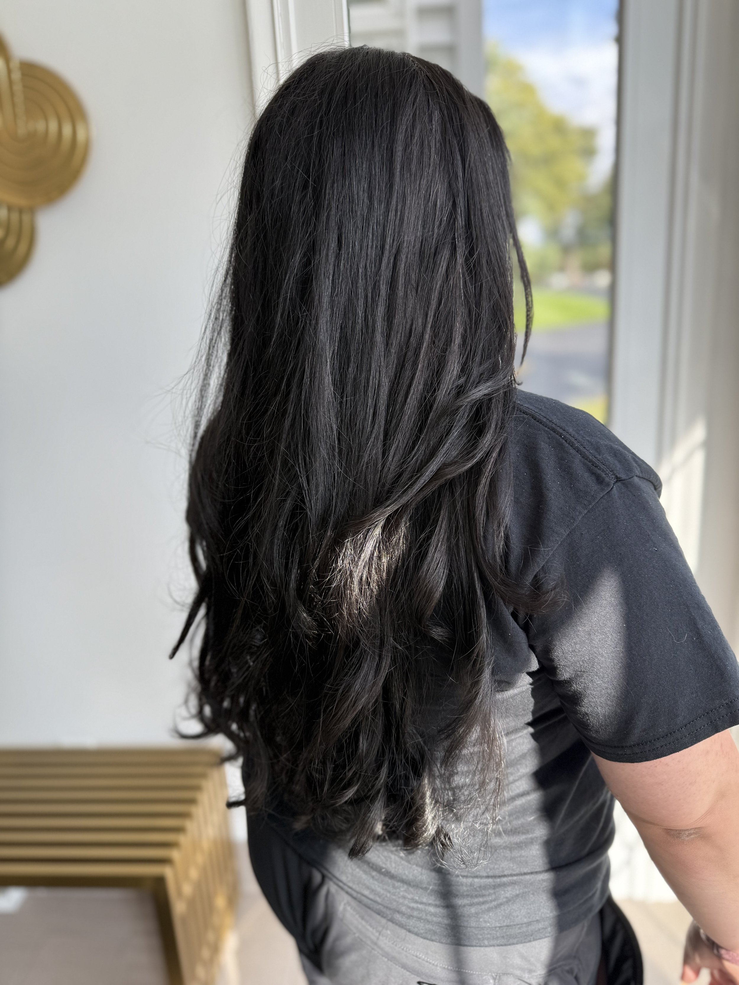 Person with long, dark hair wearing a black t-shirt and light gray sweatpants, captured from behind, indoors near a window with a view of trees and sky.