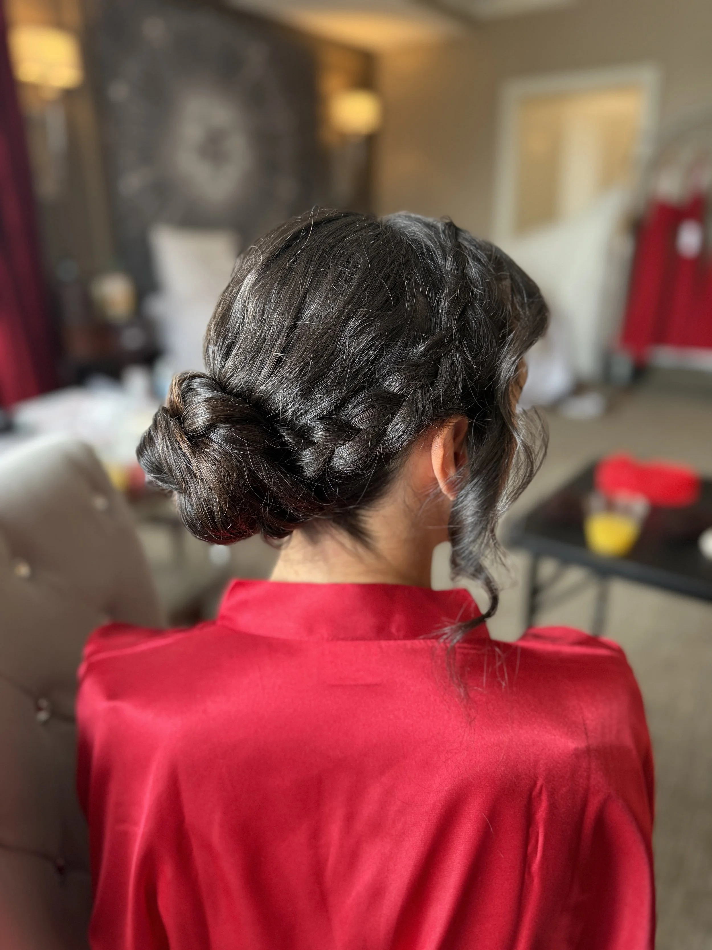 Back view of a woman with dark hair styled in a loose braid, wearing a red sweater, in a room with blurred background.
