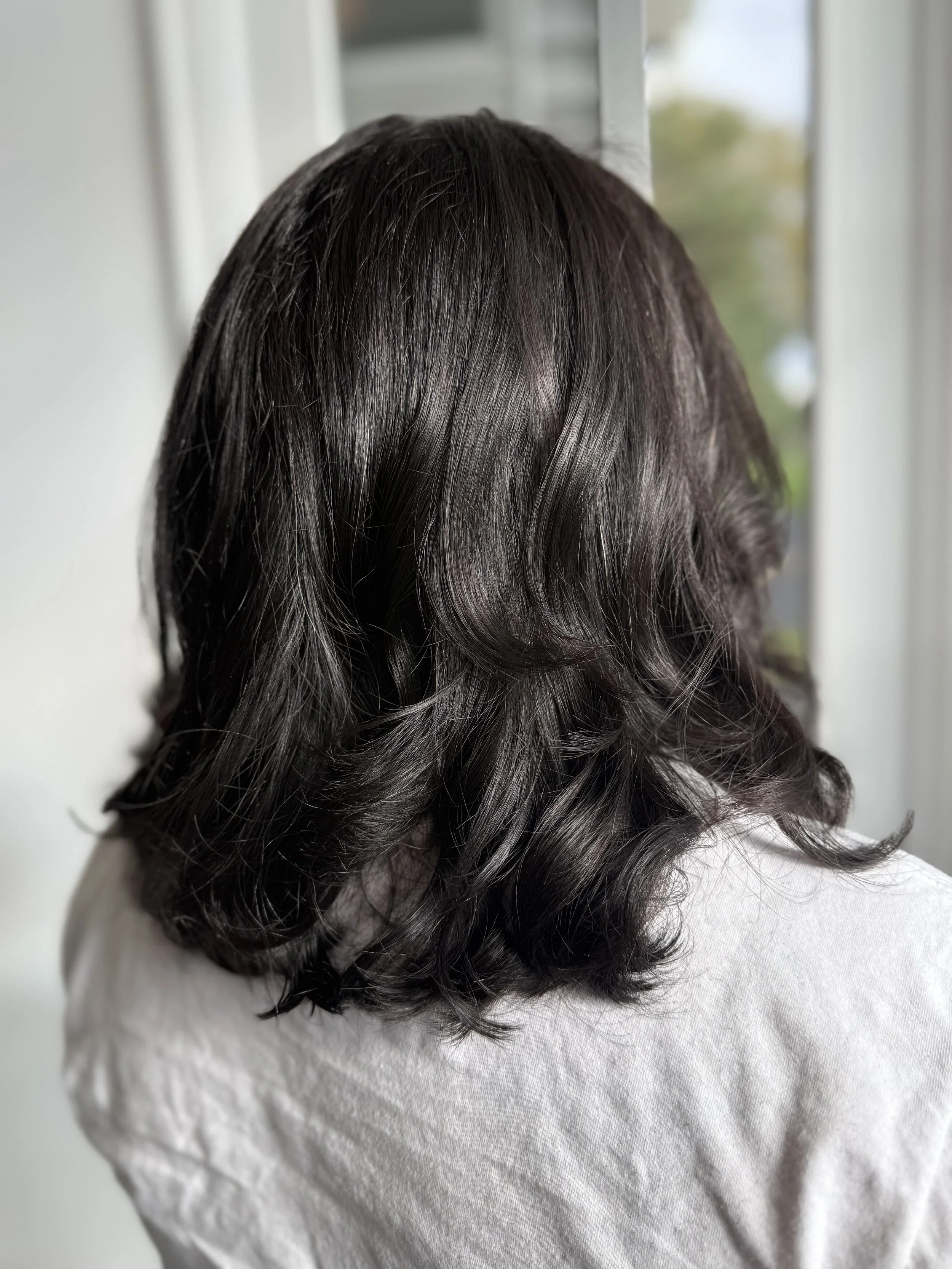 Close-up of dark, wavy, medium-length hair on a person's head, standing on a white textured surface, with a blurred background.