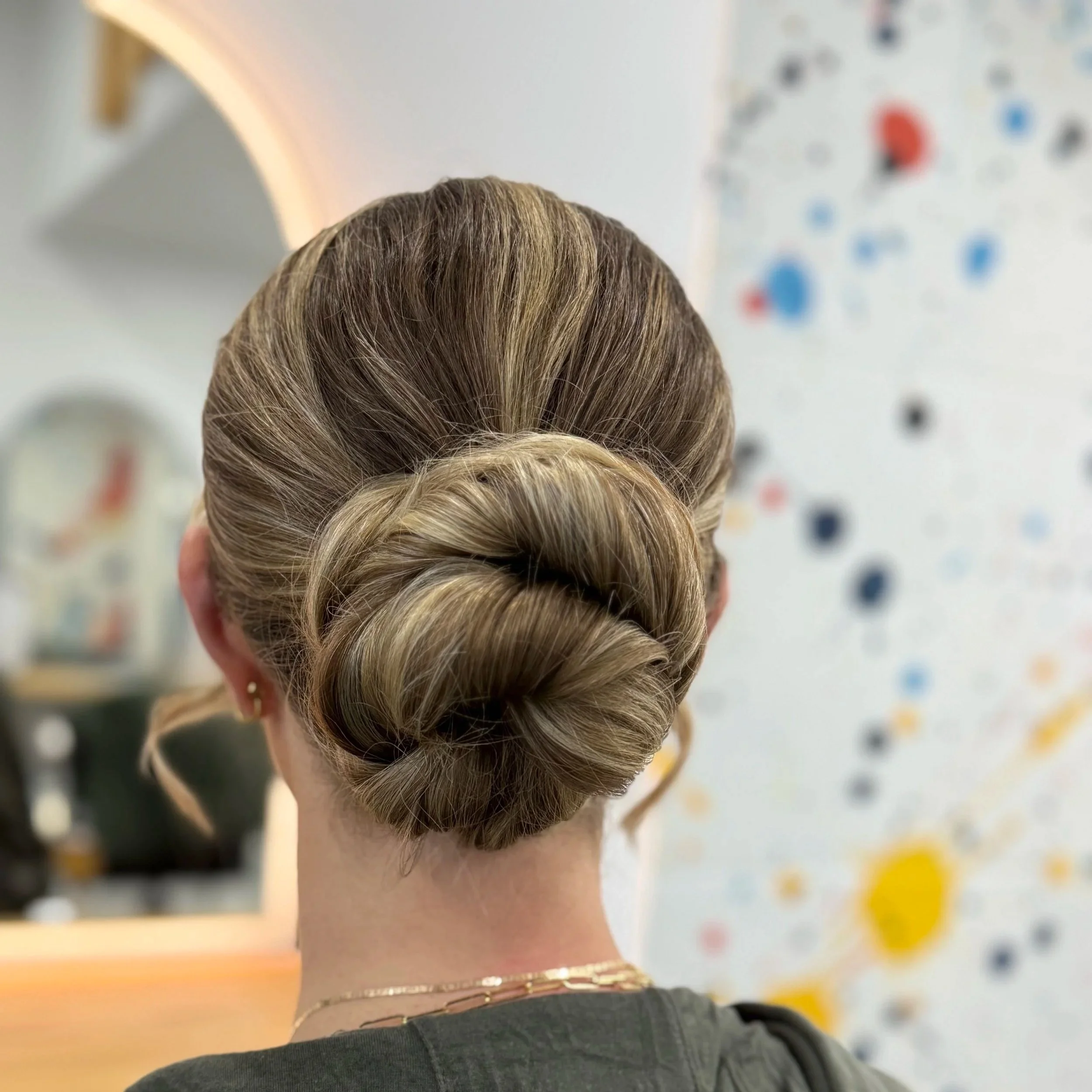 Back view of a woman with a braided bun hairstyle, wearing a black top, gold chain necklace, and gold earrings, in an indoor setting with colorful blurred background.