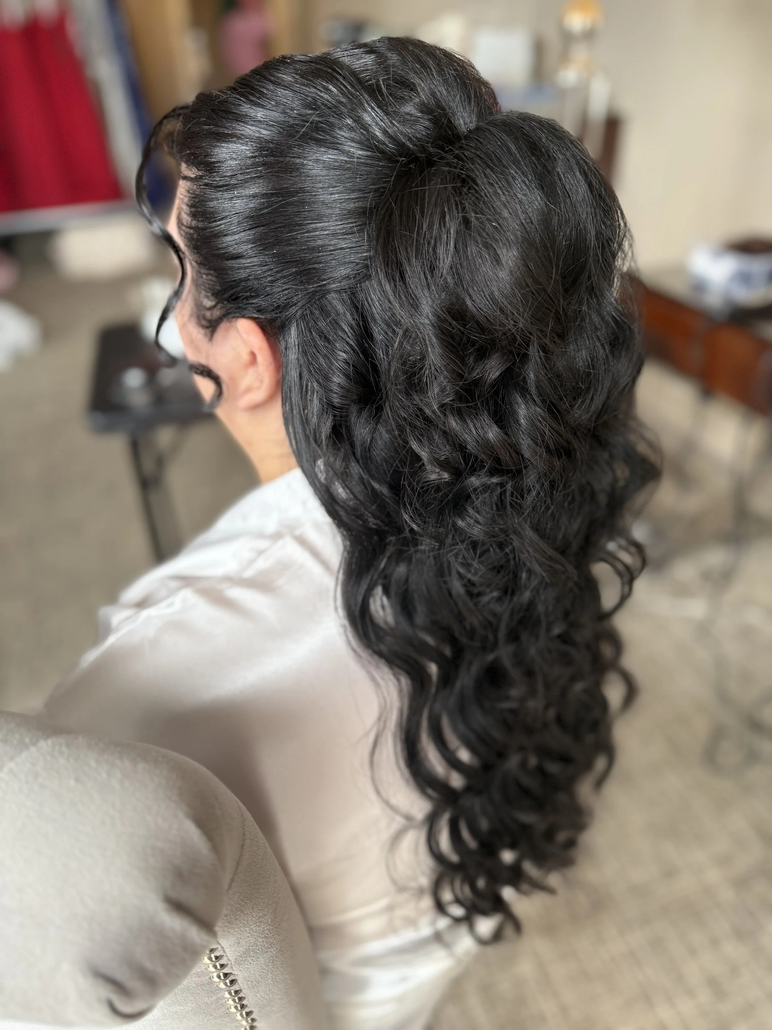 A woman with long, dark, curly hair. She is wearing a light-colored satin or silk top or dress, with a metallic zipper detail along the sleeve. The background shows a blurred room with some furniture and objects.