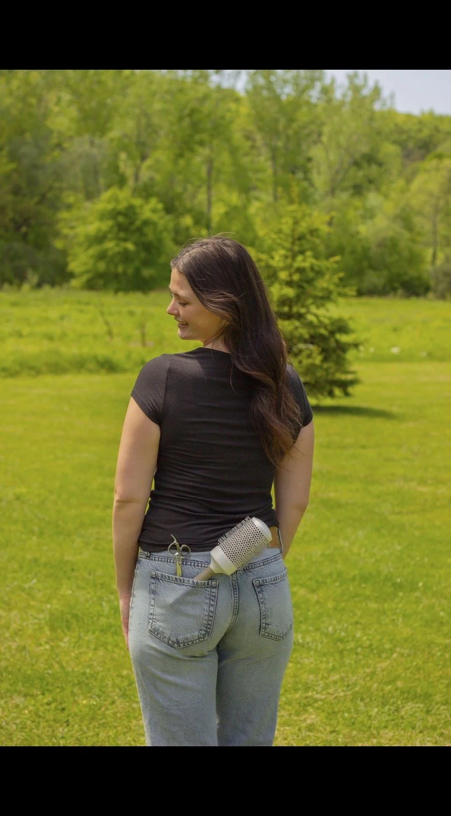 Young woman standing outdoors on a grassy field, with her back facing the camera, holding hair cutting scissors and a hairdryer in her back pocket, wearing a black t-shirt and light blue jeans, surrounded by green trees and foliage.
