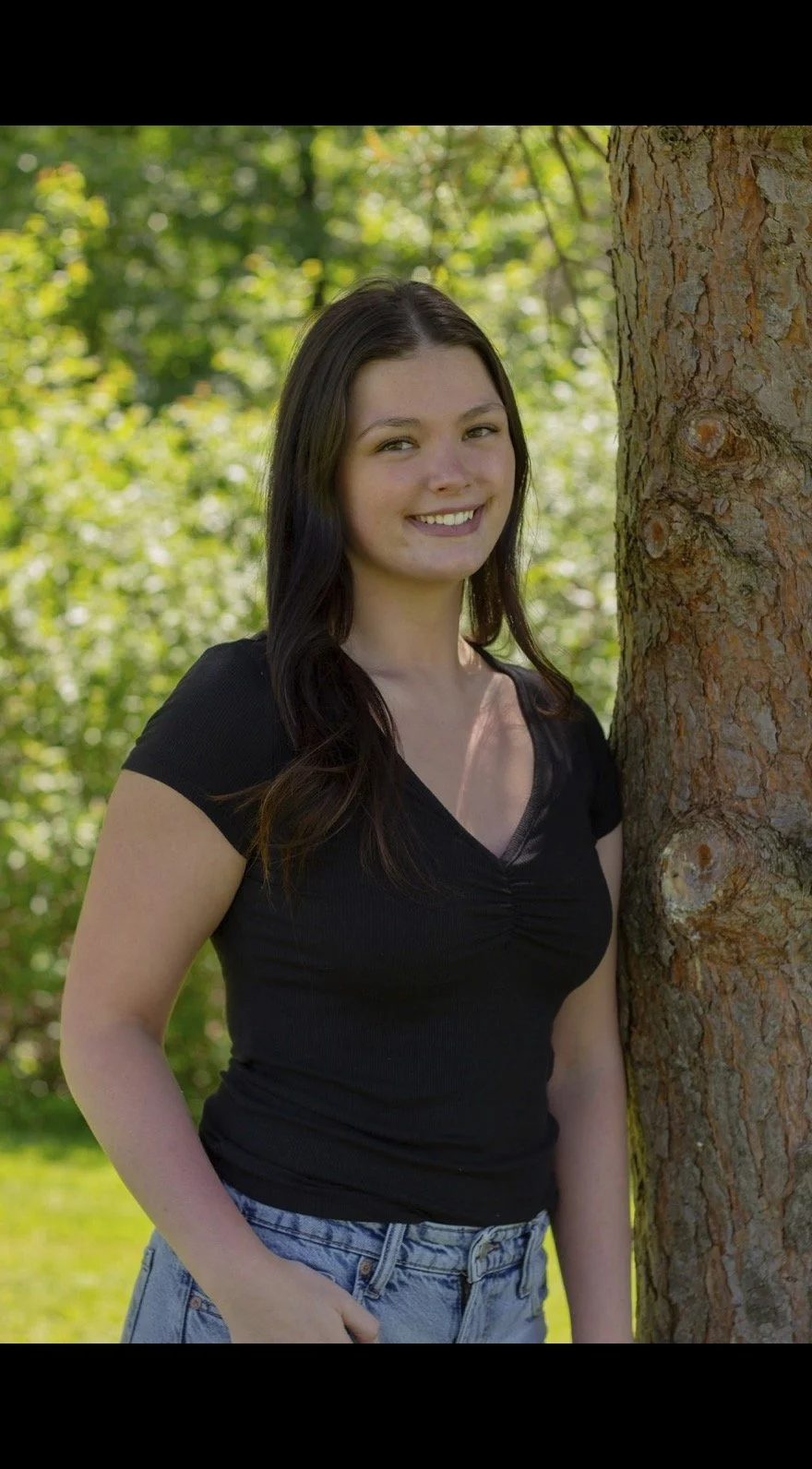 Young woman with long dark hair smiling and standing outdoors next to a tree, wearing a black top and blue jeans, with a background of green foliage.