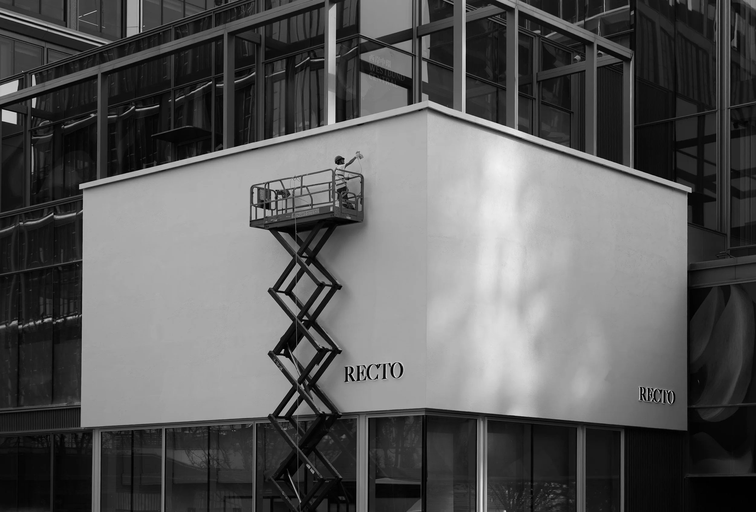A worker on a scissor lift cleaning the exterior glass windows of a modern multi-story building with the word 'RECTO' visible on its facade in black uppercase letters.