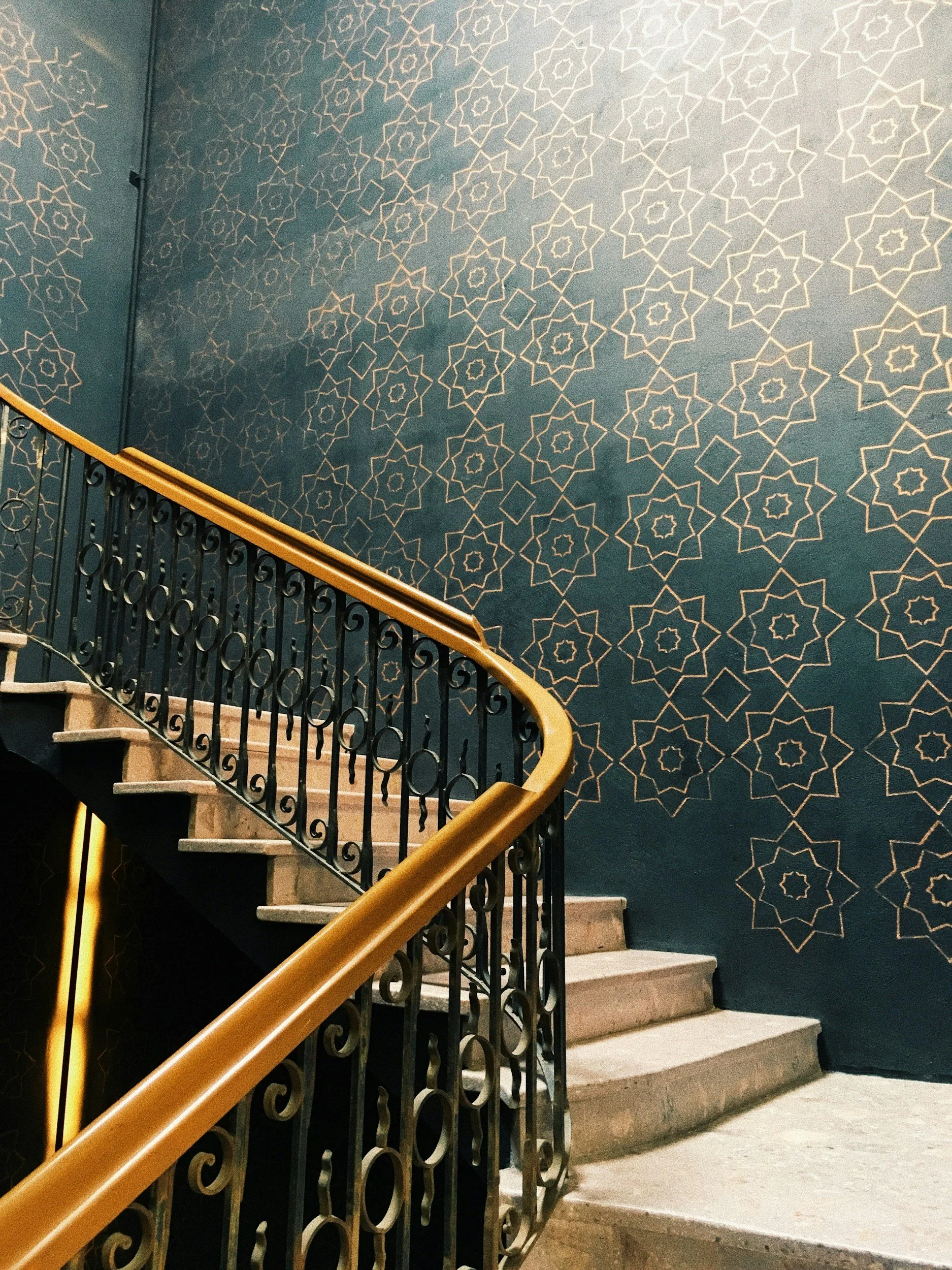 Interior staircase with ornate black metal railing and a wooden handrail, set against a blue wall decorated with a pattern of interconnected geometric shapes in a star and diamond design.