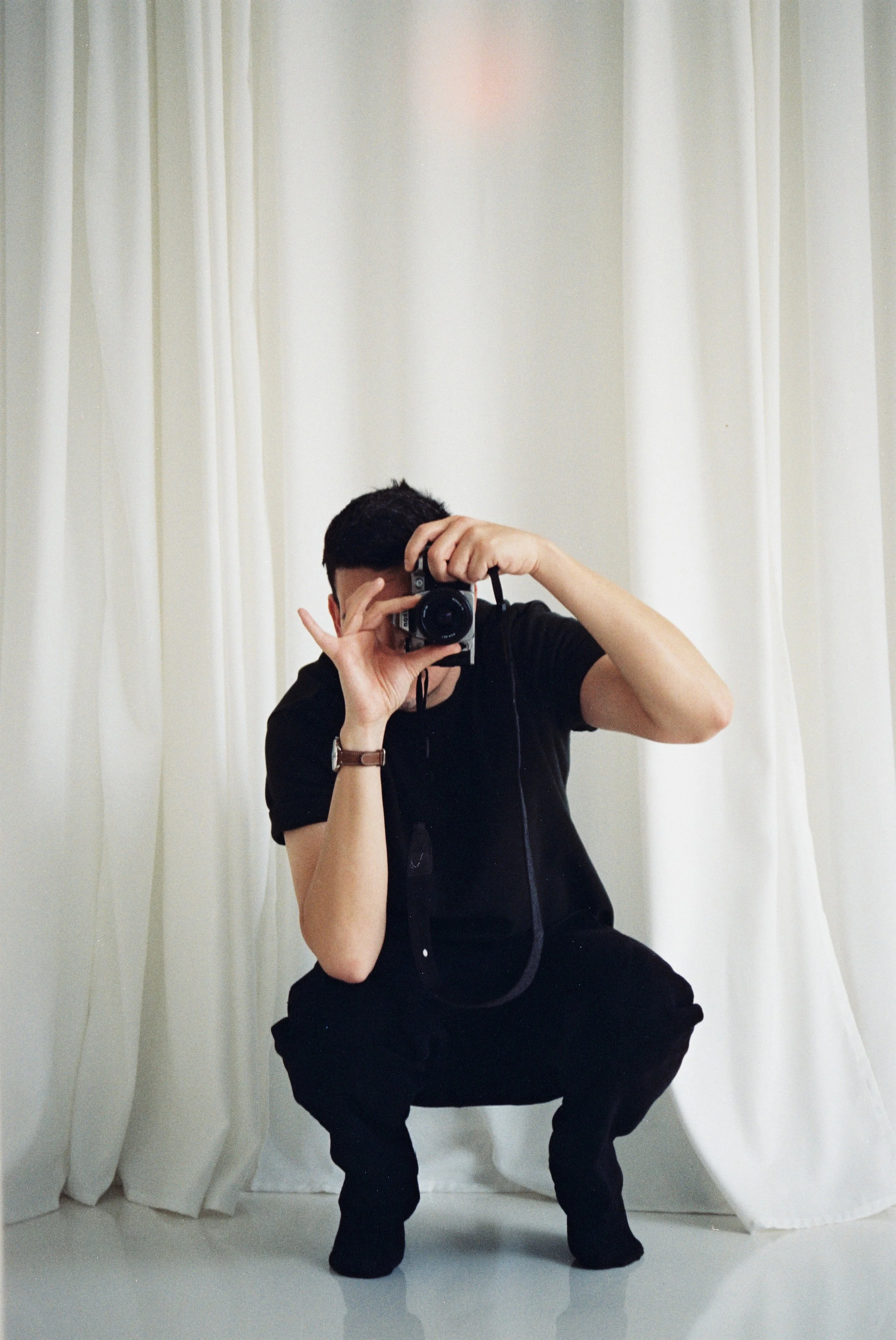 Person with dark hair wearing black clothing and a bracelet, taking a photo with a camera in front of a mirror with white curtains, squatting down.