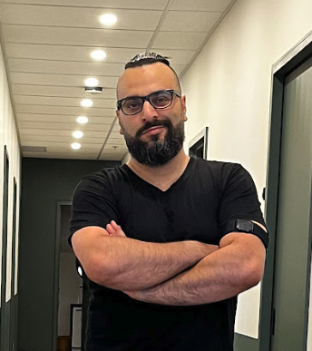Homme avec barbe et lunettes, portant un t-shirt noir, croisant les bras, dans un couloir intérieur.