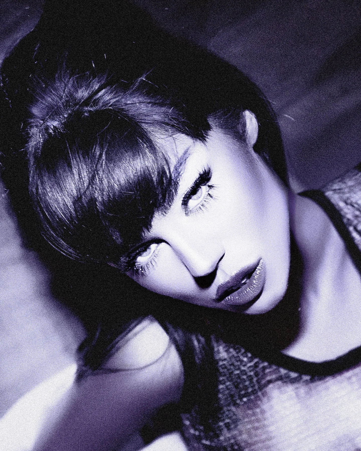 A close-up, black-and-white photograph of a woman's face with her eyes closed, showing detailed makeup and long eyelashes, with dark hair and textured clothing.