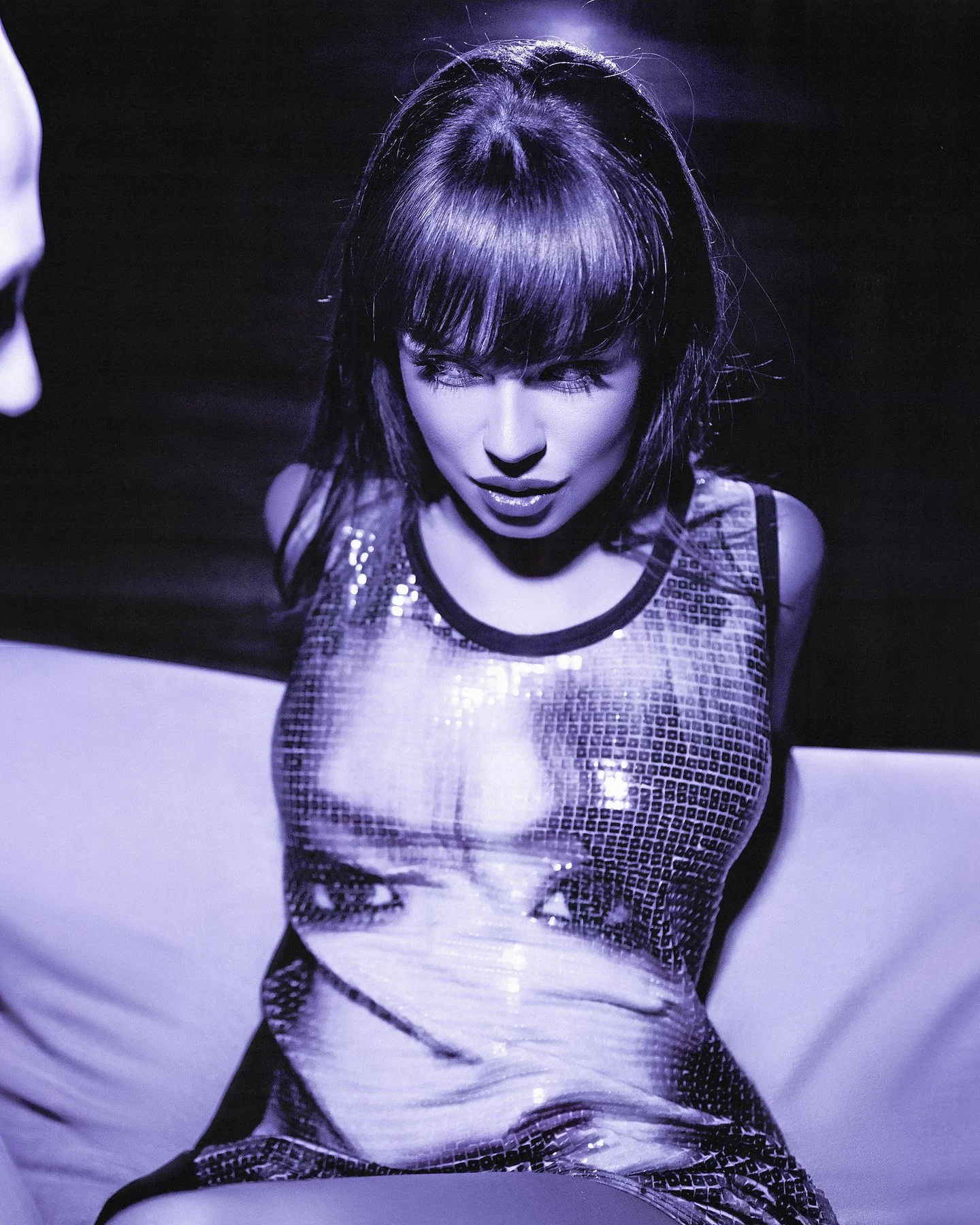 A woman with dark hair and bangs, wearing a shiny, reflective, metallic top, looking downward with a slightly open mouth.