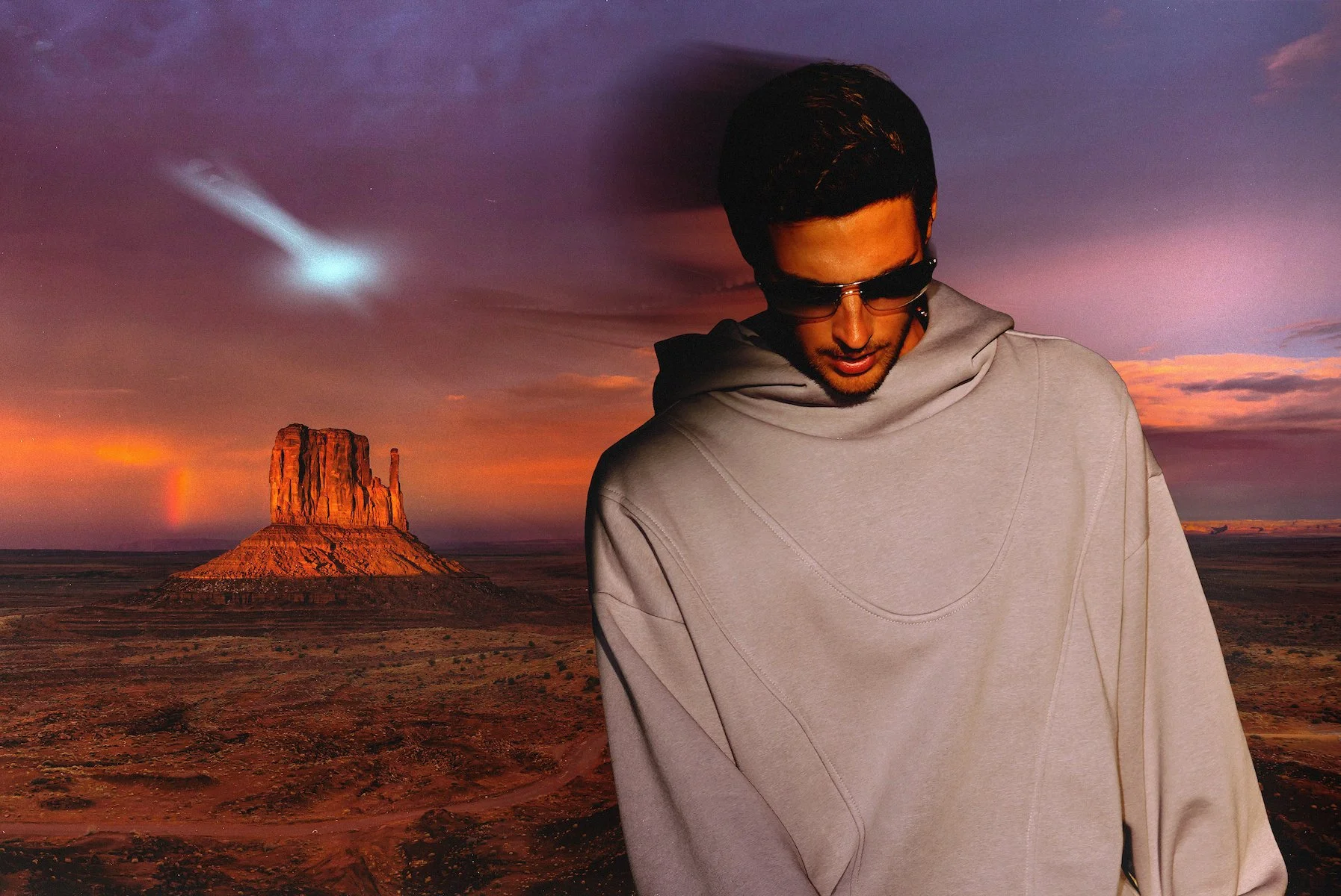 A young man wearing sunglasses and a light gray hoodie stands in front of a desert landscape with a large butte and colorful sunset sky.