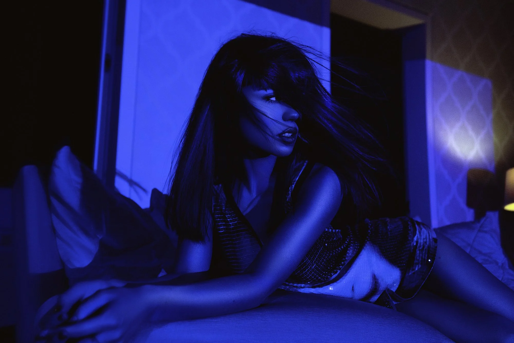 A woman with long hair sitting on a bed in a dimly lit room illuminated by blue light.