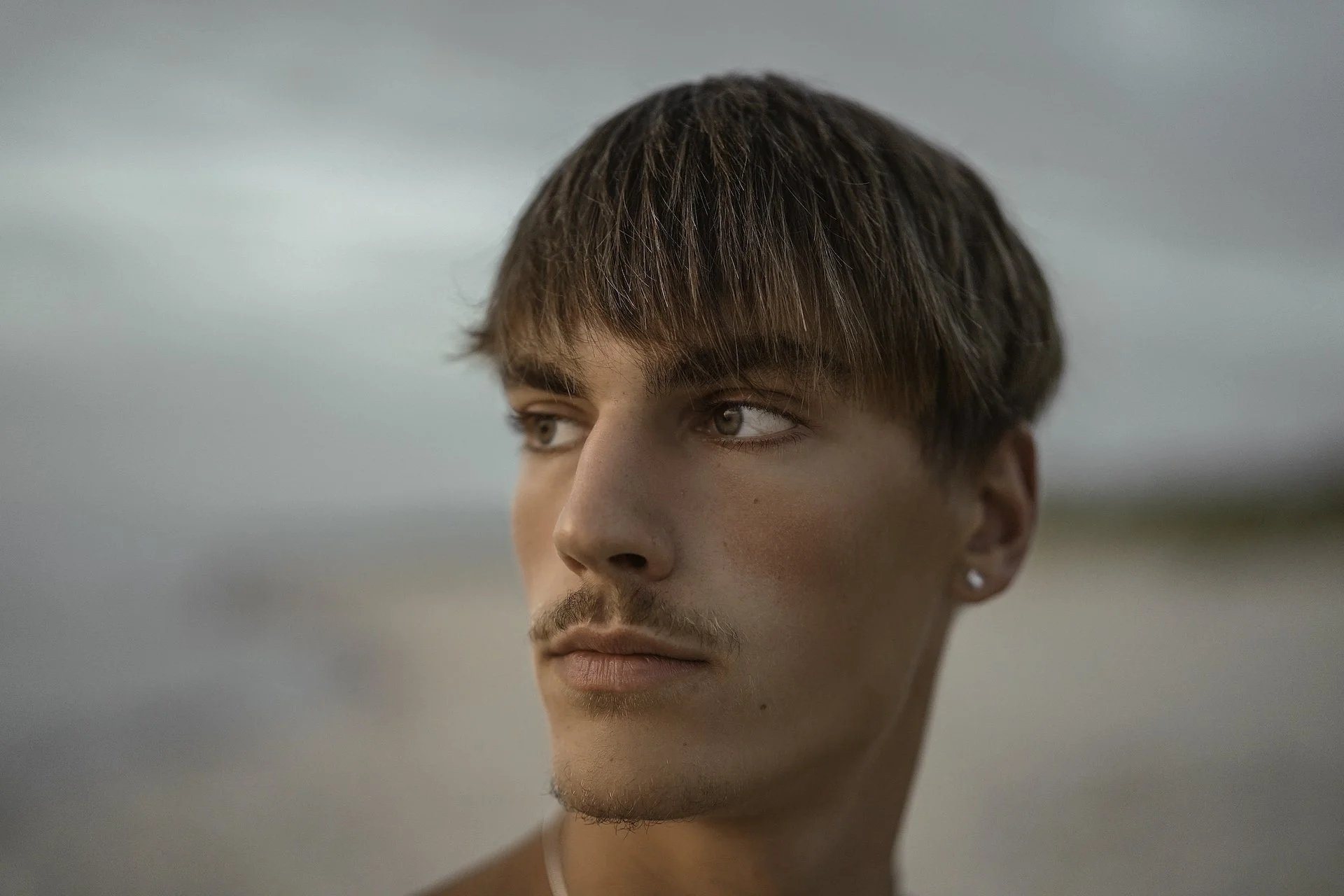 Close-up of a young man with short brown hair, brown eyes, and earrings, looking to the side against a blurred outdoor background.