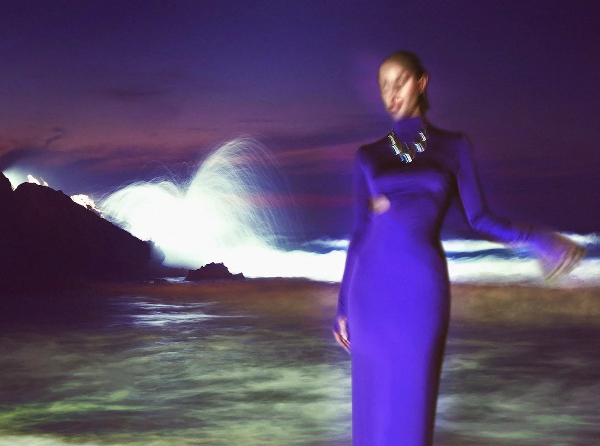 A woman in a long purple dress standing on a beach at night, with large waves crashing behind her, and water spray illuminated by the wave.