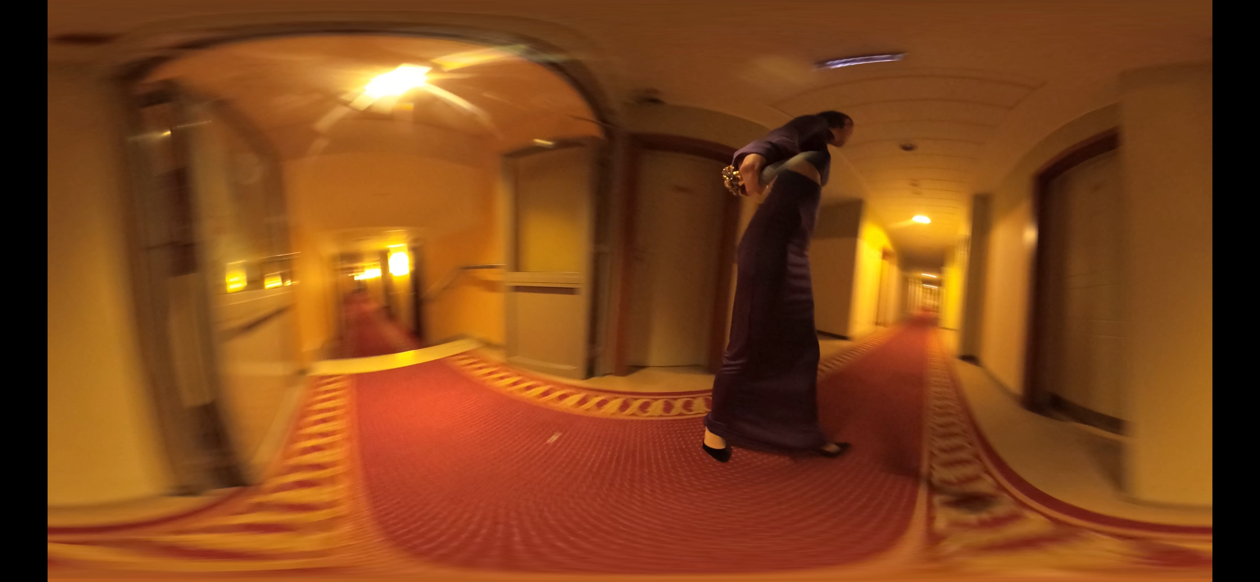 A woman in a black dress walking down a hotel hallway with a red carpet and yellow walls, holding a handbag.