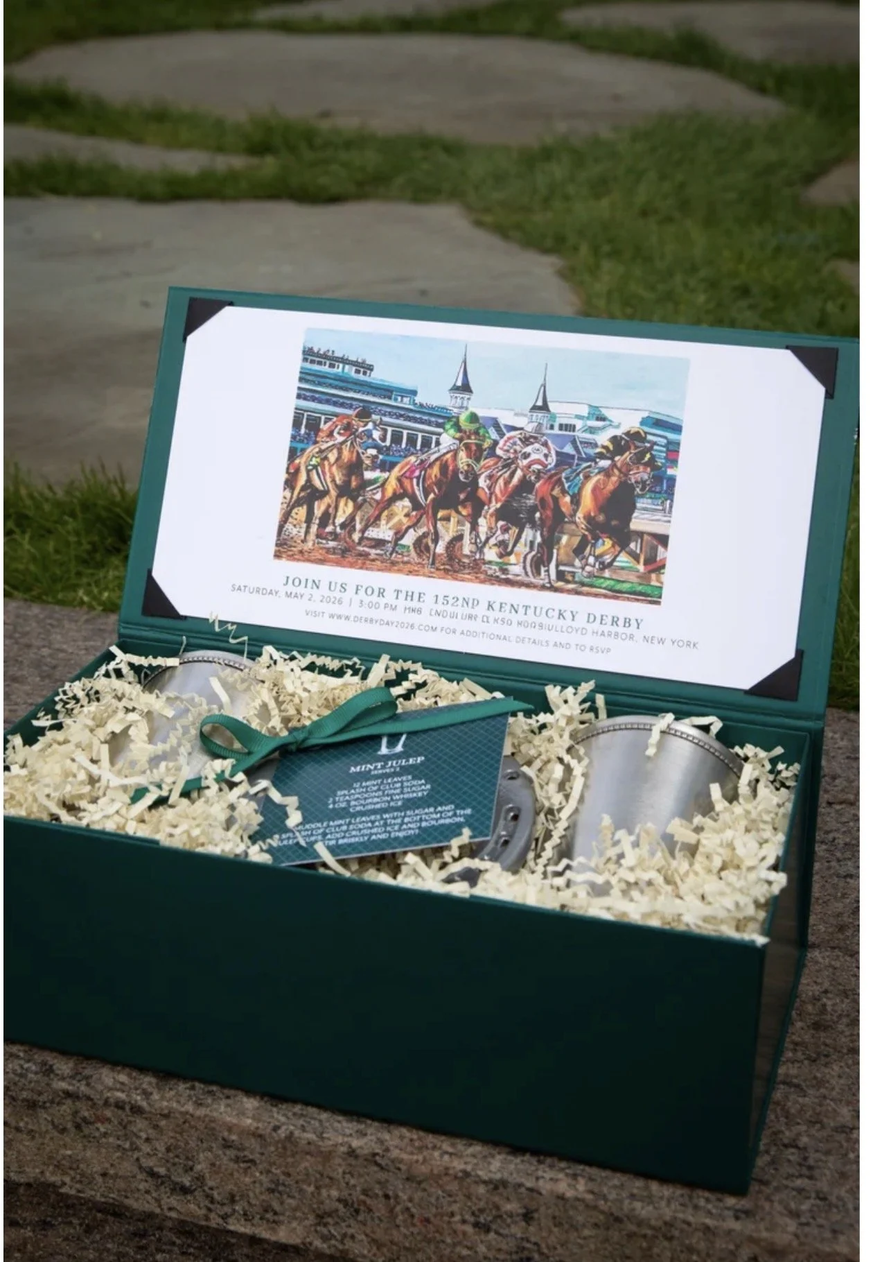A teal gift box with shredded paper, two silver bracelets, and a card inside. The card features an illustration of horse racing and text inviting to a Kentucky Derby event around a horse race theme.
