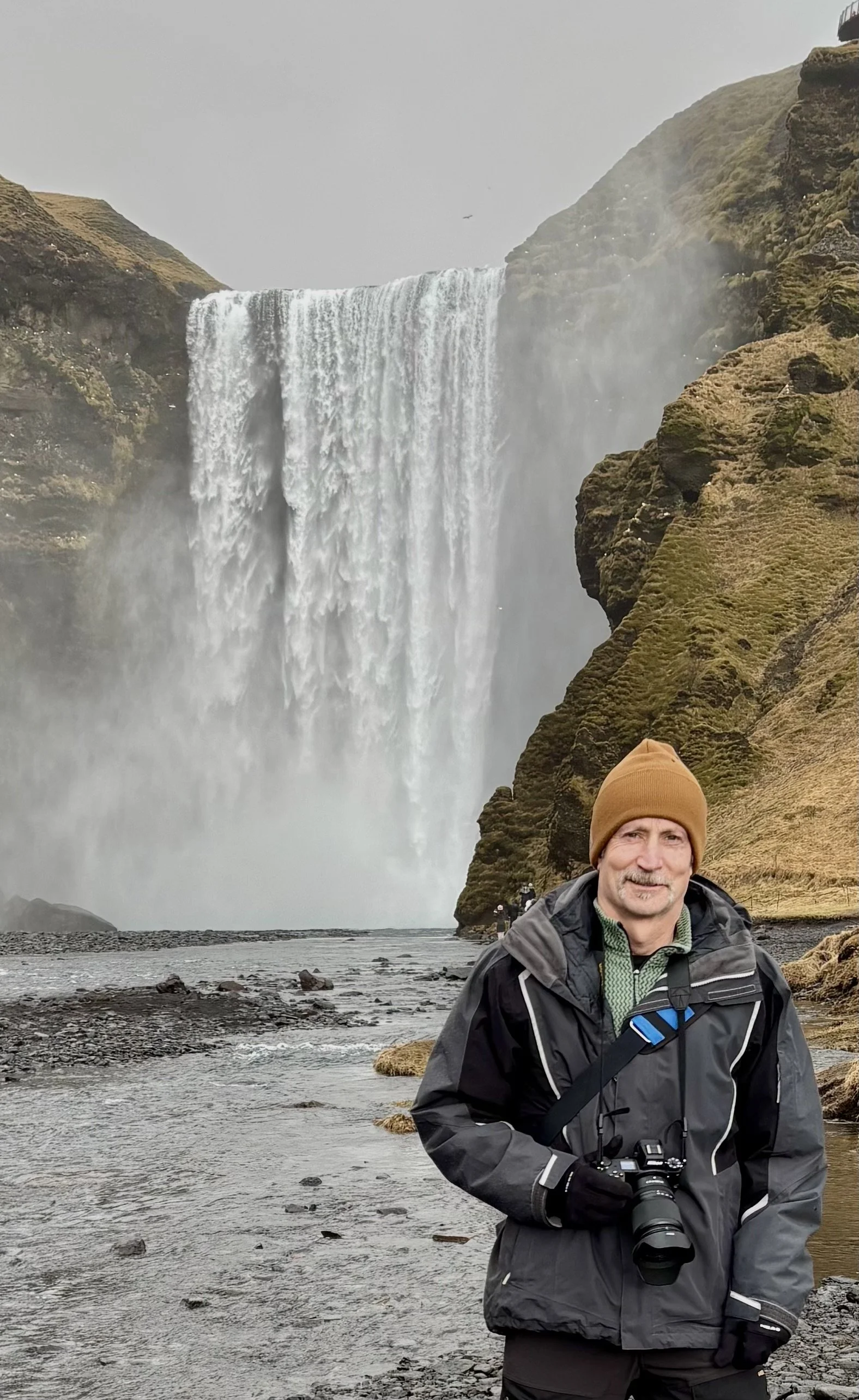 The author stands in front of Skogafoss waterfall in Iceland, with rocky terrain and moss-covered cliffs in the background. He is wearing a warm jacket and brown beanie to ward off the cold, and is holding a long lens camera