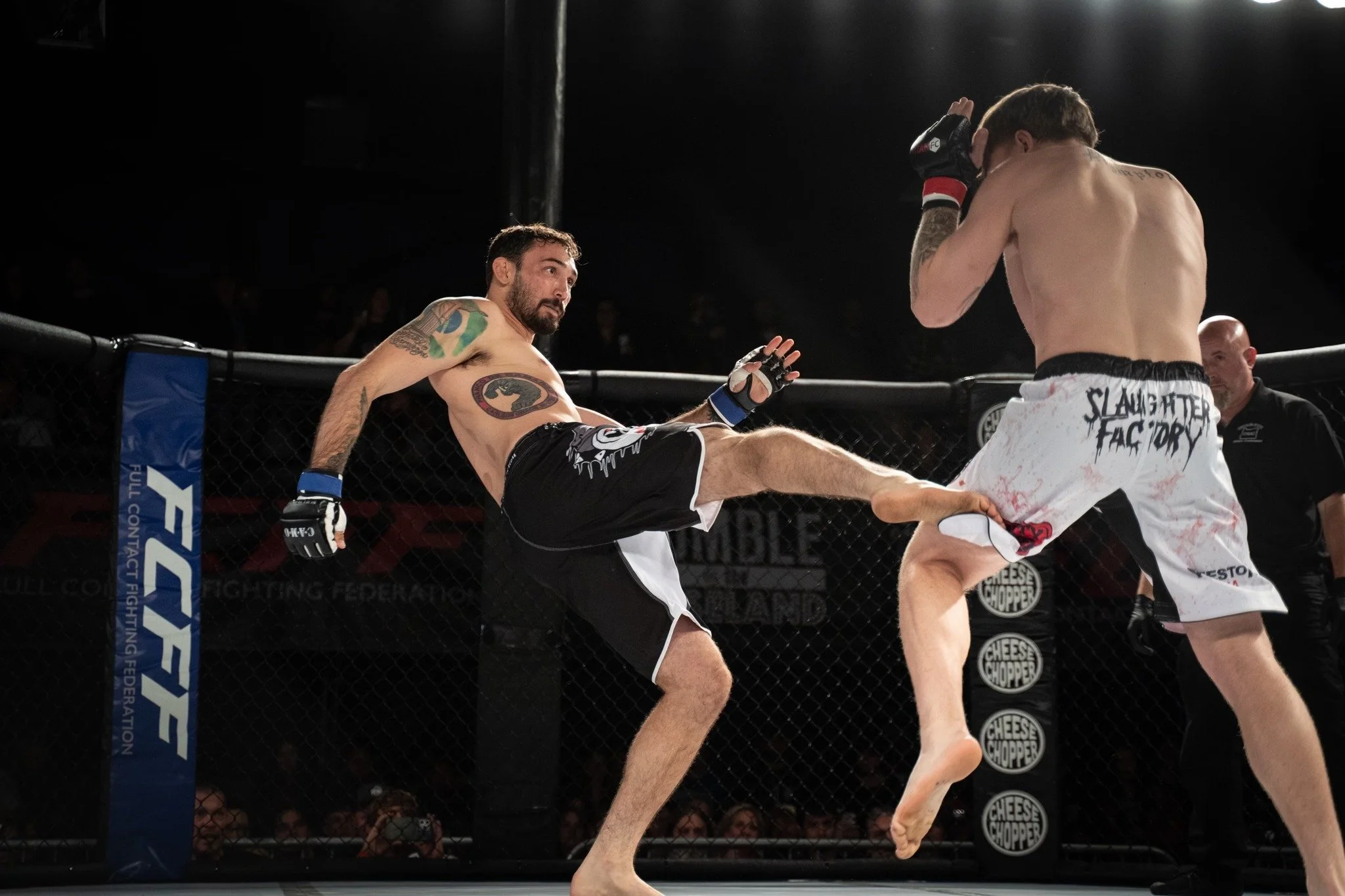 Two mixed martial arts fighters are engaged inside an octagon cage. One fighter, with tattoos and wearing black shorts, is delivering a high kick to the other fighter, who is wearing white shorts. The fighter in white shorts is shielding his face with his hands. The background shows the octagon’s fence and a dark arena setting.