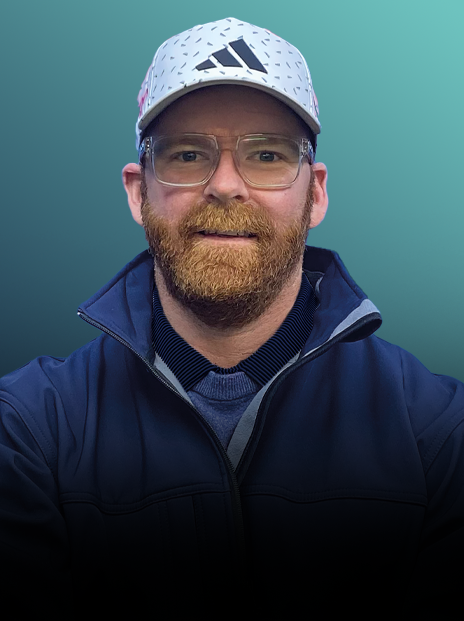 A man with glasses, a beard, and a mustache wearing a white athletic cap with black logos, a dark zip-up jacket, and an indigo shirt, taking a selfie with a teal background.
