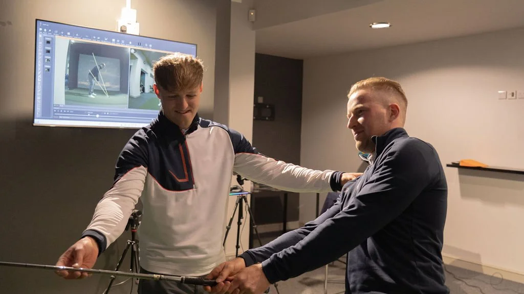 Two men in a room with a golf simulation screen, one adjusting the other's arm for a golf swing analysis.