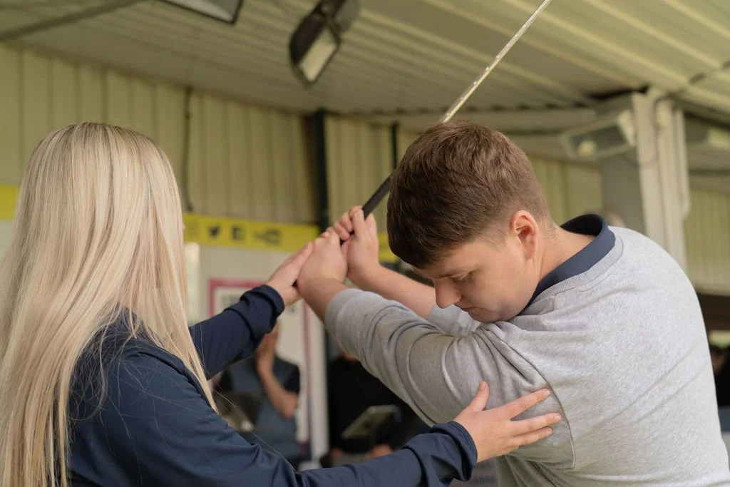 A woman assists a young man in stretching with a golf club tool in an indoor gym or training area.