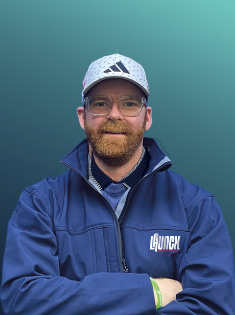 A man with glasses and a beard wearing a blue jacket and a white cap, standing with arms crossed against a gradient background.