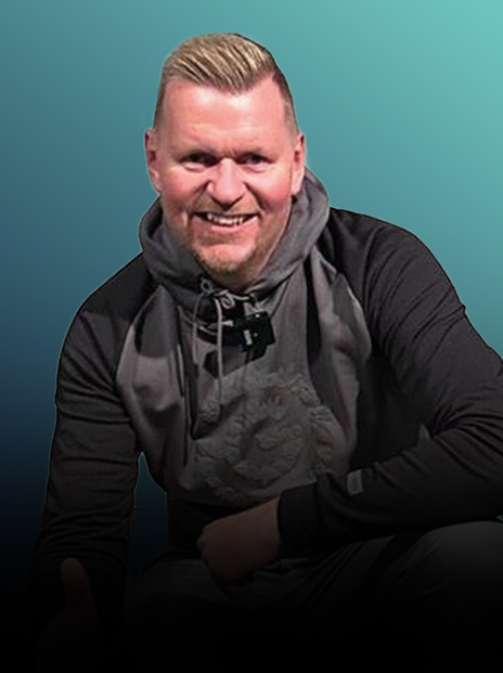 A smiling man with short blond hair, beard, and wearing a black and gray hoodie, sitting against a gradient teal background.
