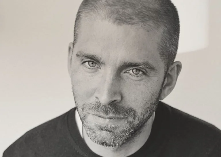 Close-up black and white portrait of a man with short hair and beard, looking directly at the camera.
