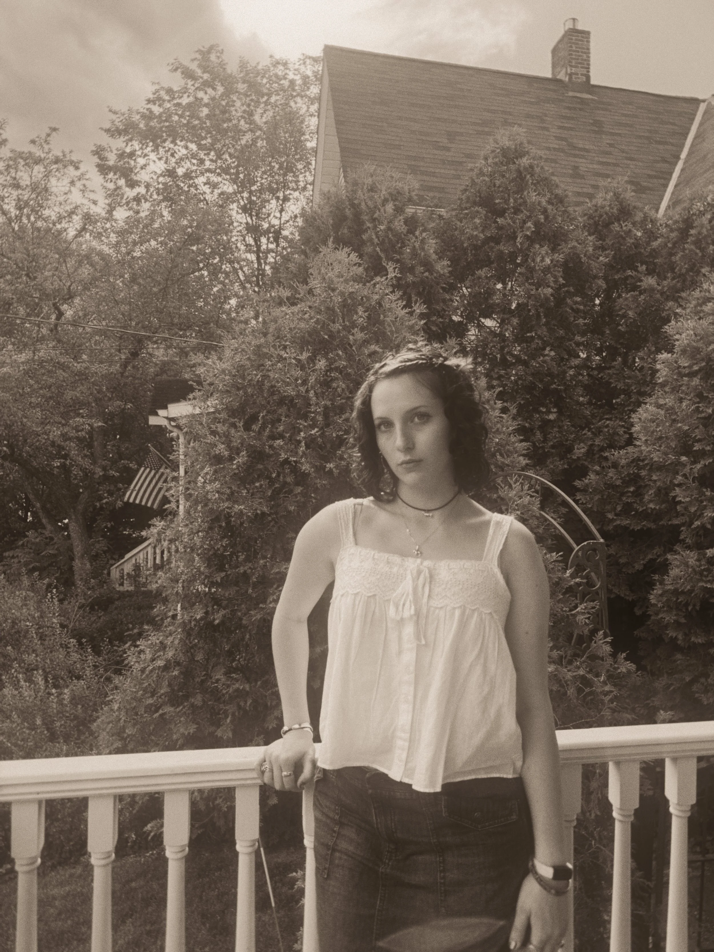 A woman standing outdoors on a porch, leaning on a railing, with trees and a house in the background. She has dark, shoulder-length hair, wears a sleeveless top, jeans, and a necklace.
