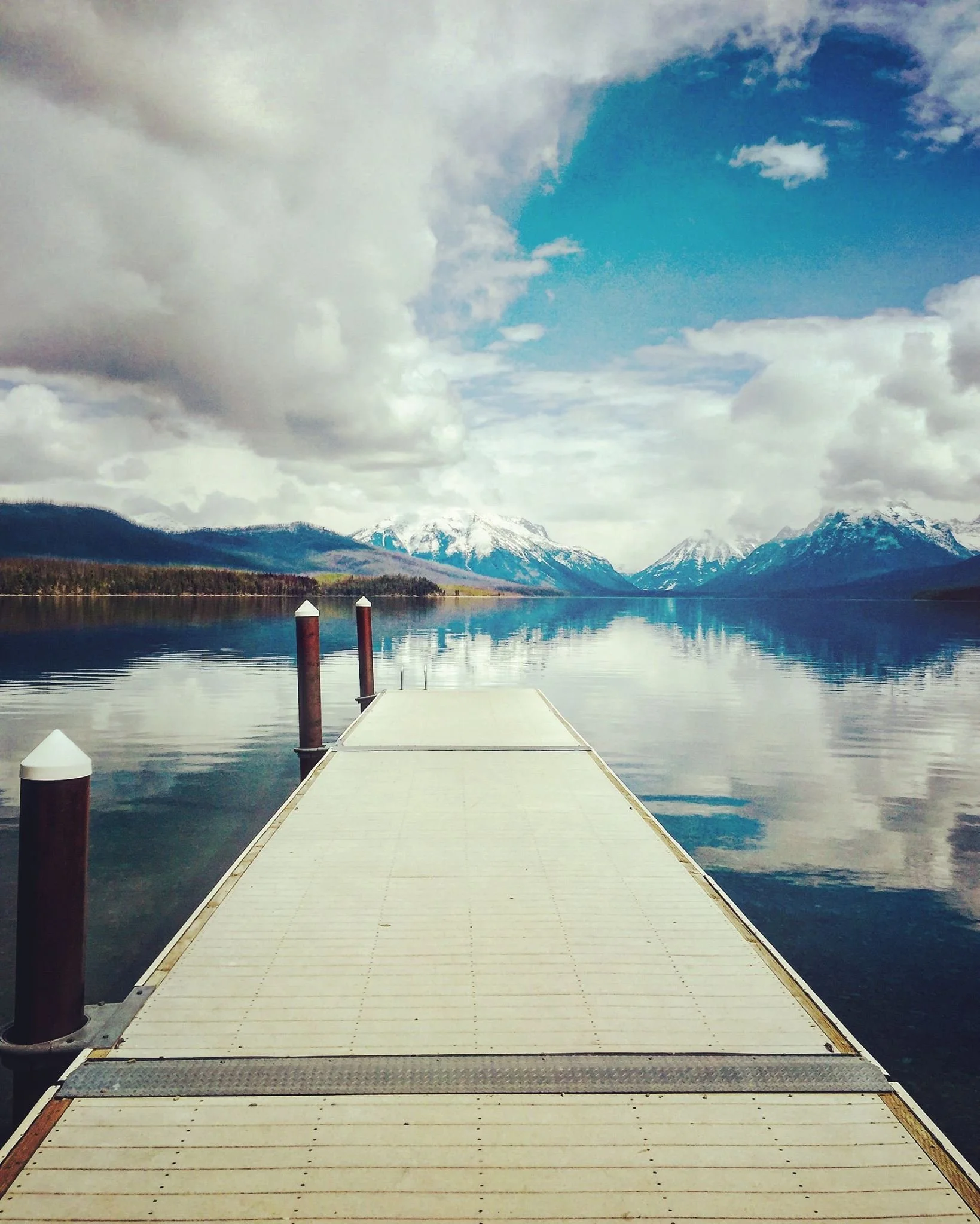 A wooden dock extends over a calm lake with snow-capped mountains and a cloudy sky in the background.