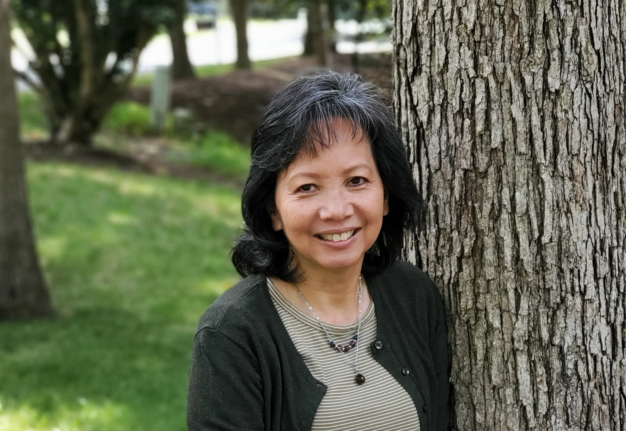 A woman with black and gray hair smiling outdoors next to a large tree trunk, with a background of green grass and trees.