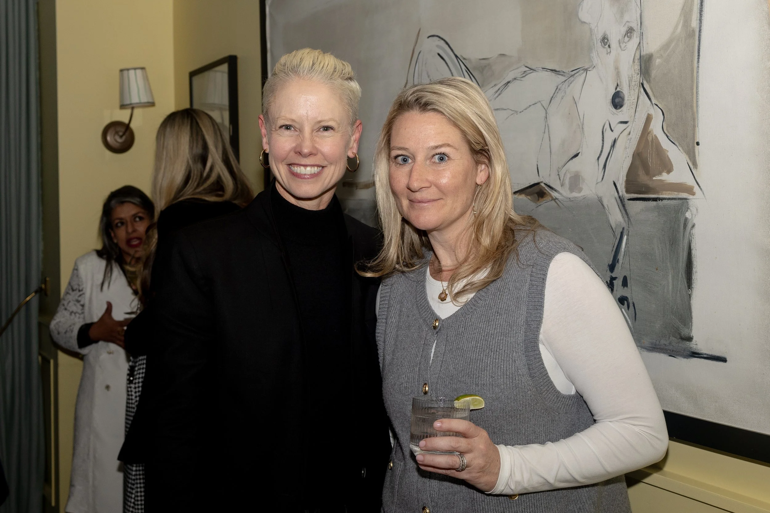 Two women standing close together at an indoor event, smiling at the camera, one holding a drink with a lime wedge, and a third woman visible in the background.