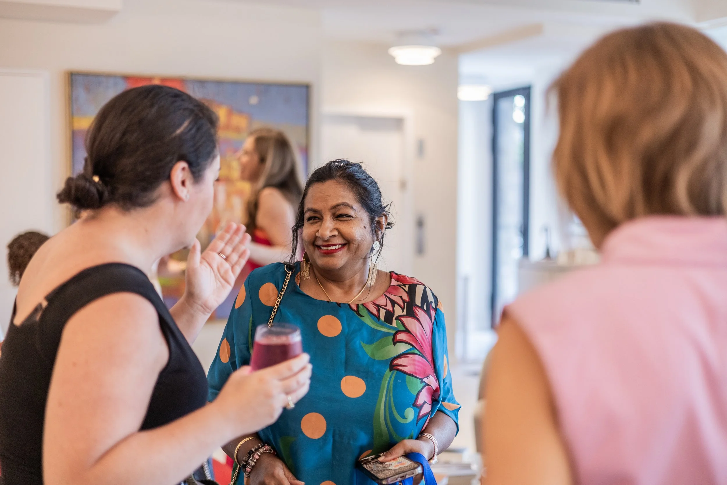Three women engaged in conversation at a social gathering indoors. One woman is holding a glass and smiling, while the other two listen and talk.