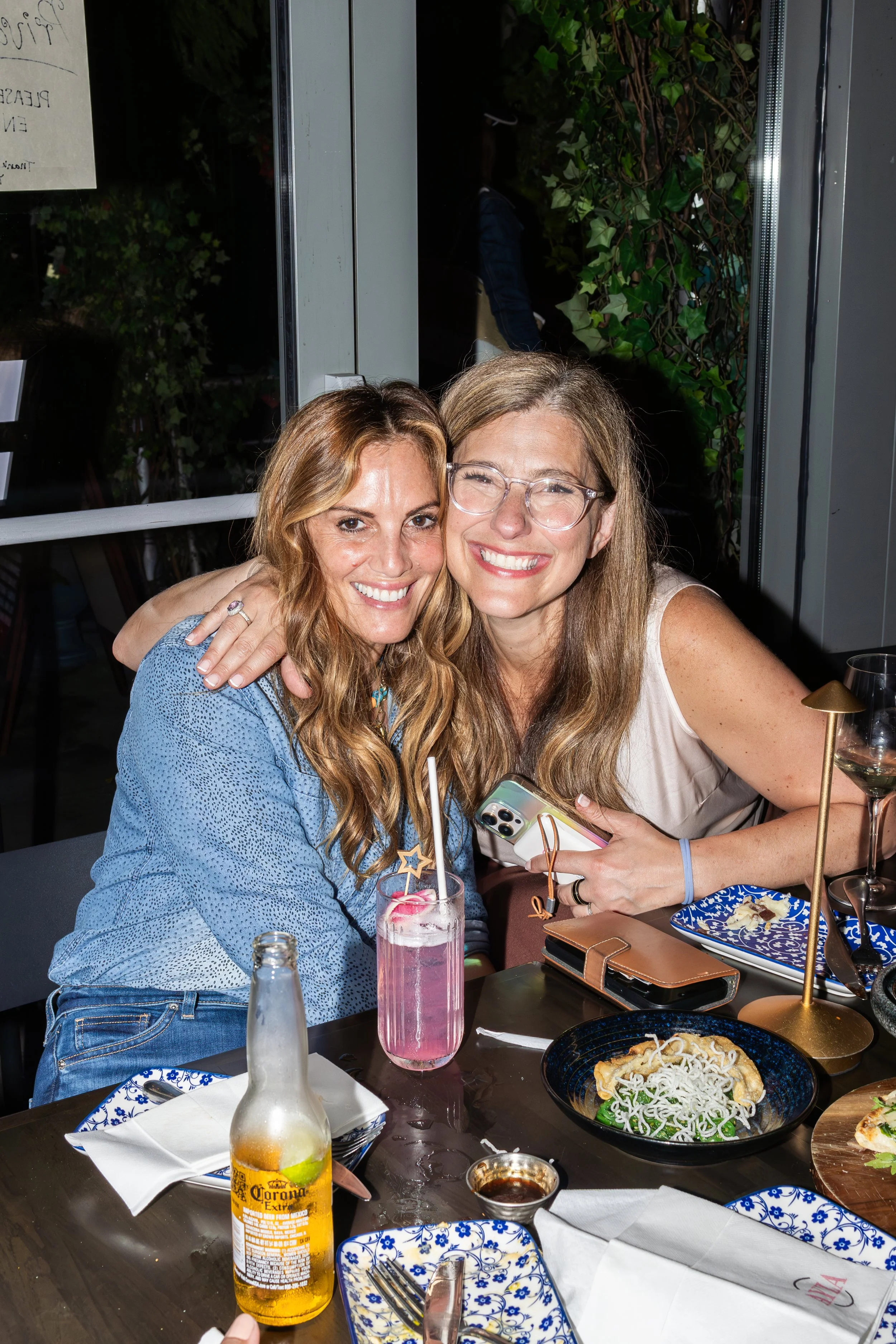 Two women smiling and hugging at a restaurant table. There are plates of food, drinks, and utensils on the table, with a window and green plants in the background.