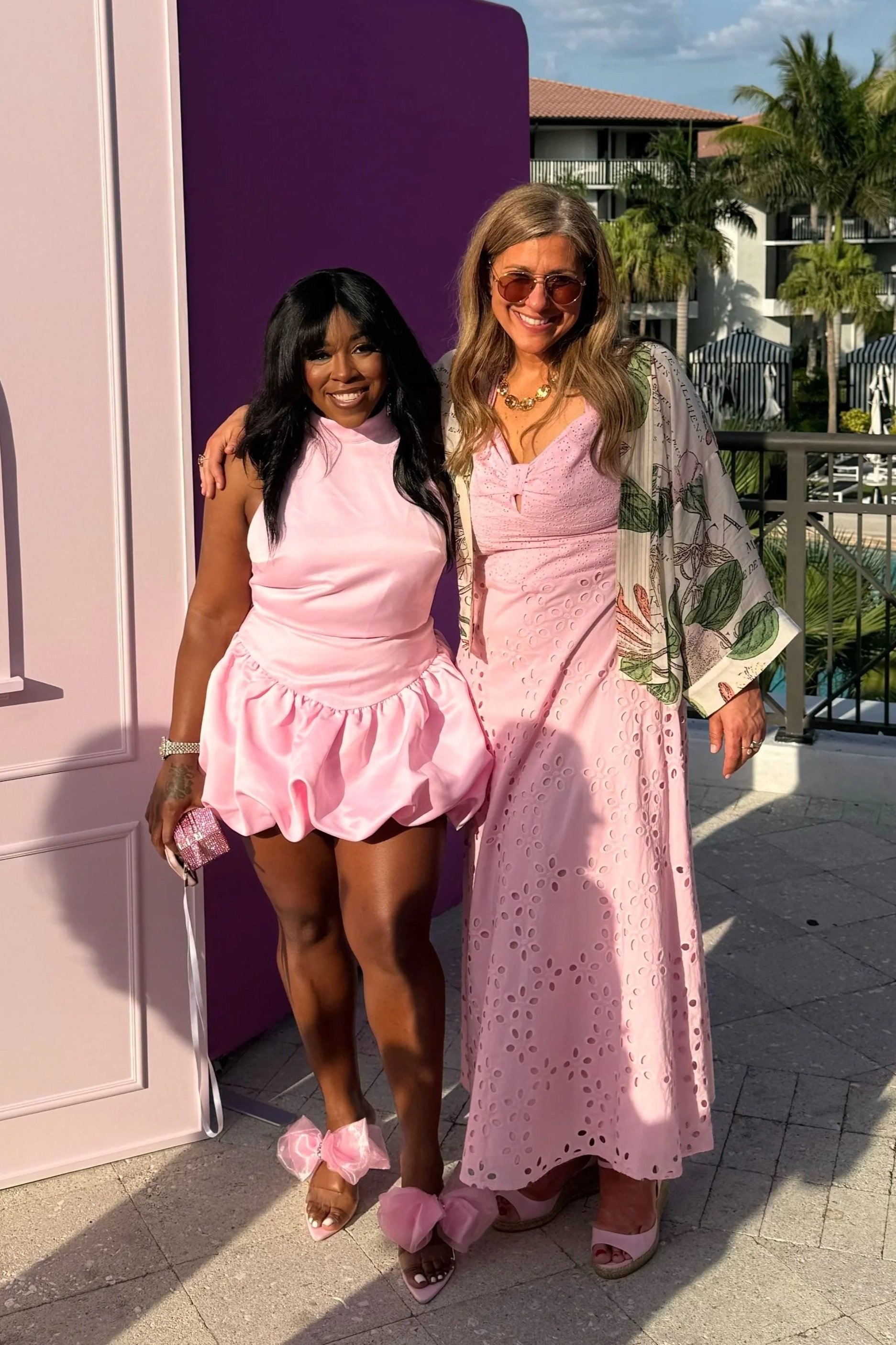 Two women standing side by side outdoors near a purple wall, dressed in pink outfits, with palm trees and a building in the background, smiling at the camera.