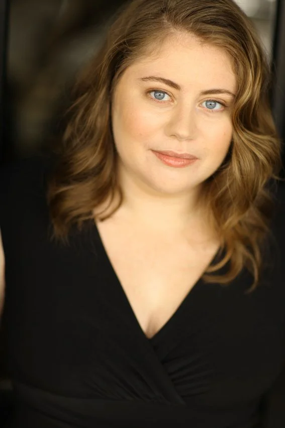 A woman with wavy, shoulder-length brown hair and blue eyes, wearing a black top, smiling softly in a close-up portrait.
