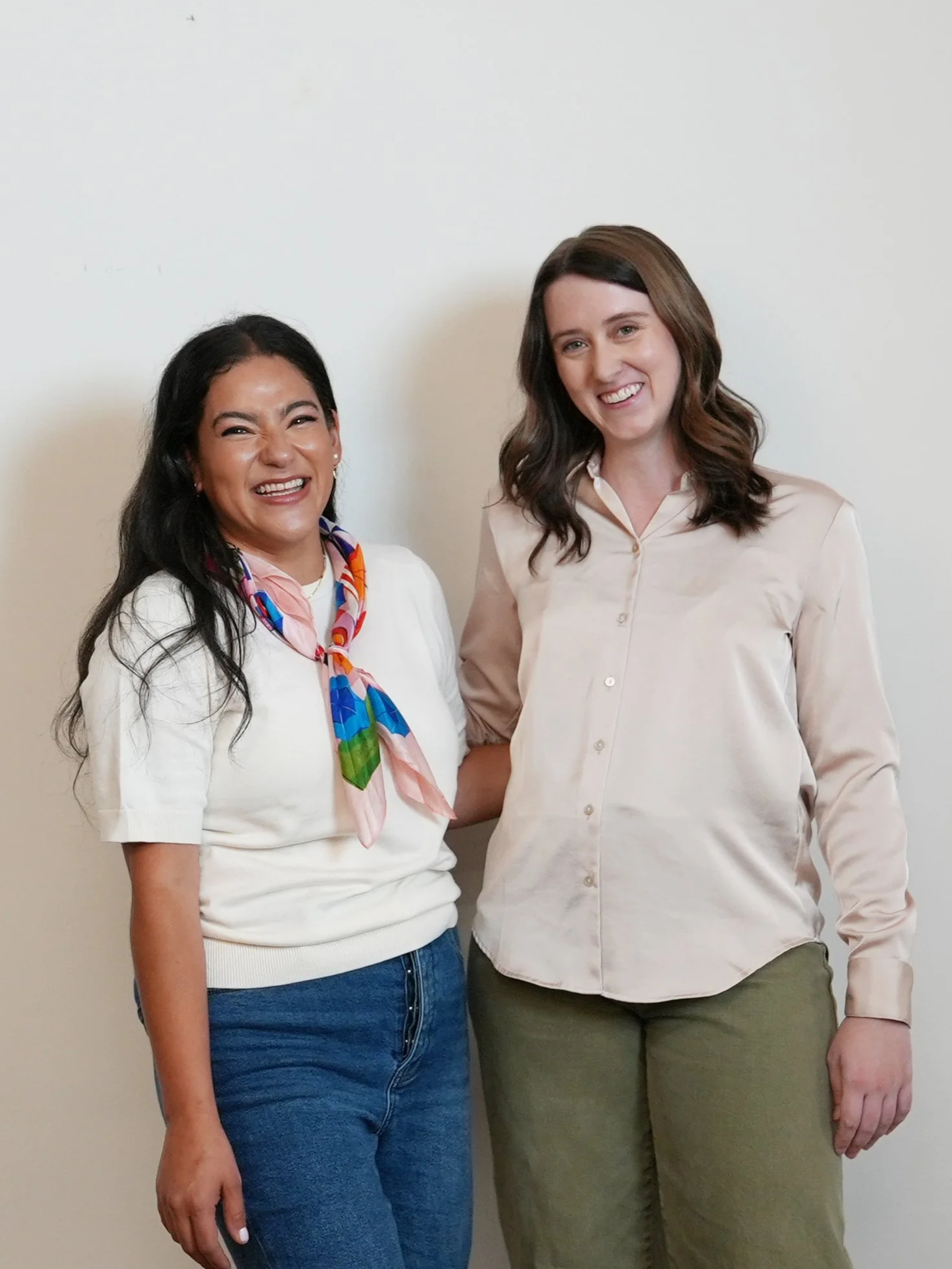 Two women standing against a plain wall smiling. The woman on the left has long dark hair, tan skin, and is wearing a white top with rolled-up sleeves, blue jeans, and a colorful scarf around her neck. The woman on the right has shoulder-length brown hair, fair skin, and is wearing a light pink button-up shirt and green pants.