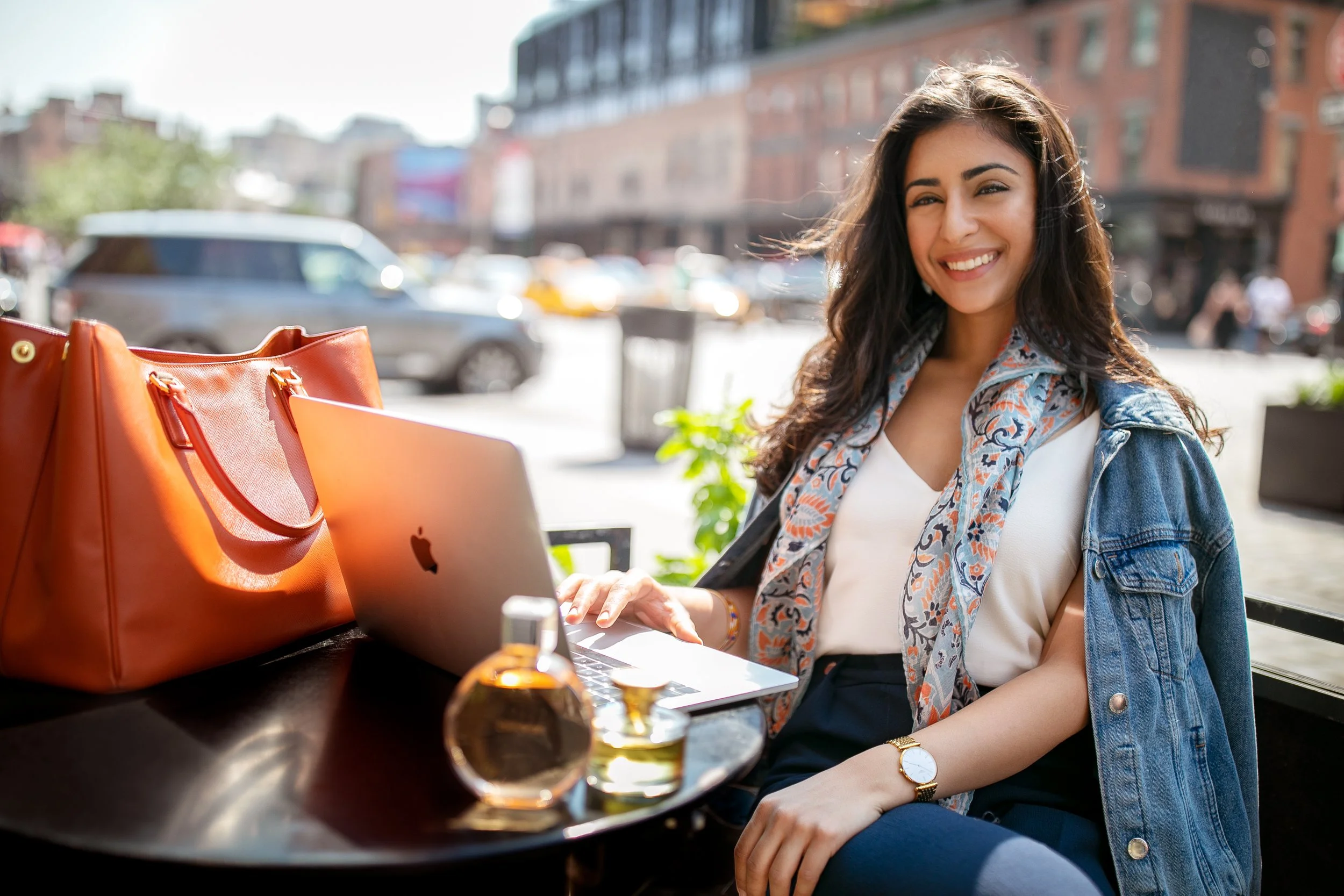 Smiling woman with dark hair, wearing a denim jacket and patterned scarf, sitting at an outdoor cafe table with a silver laptop, orange bag, and perfume bottle on table, city street with cars and buildings in background