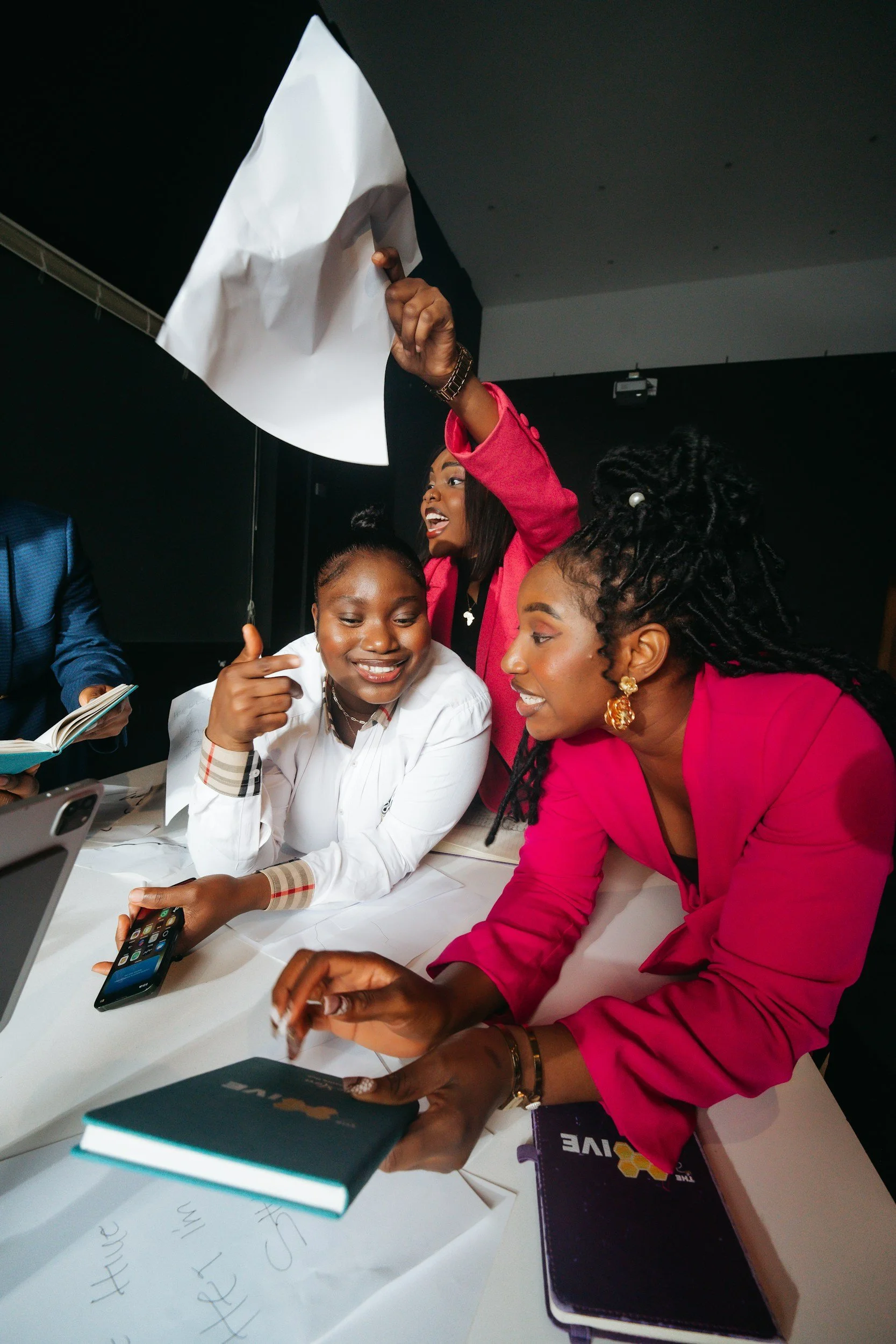 Group of women gathered around a table, taking a selfie with a smartphone, with books and notebooks on the table.