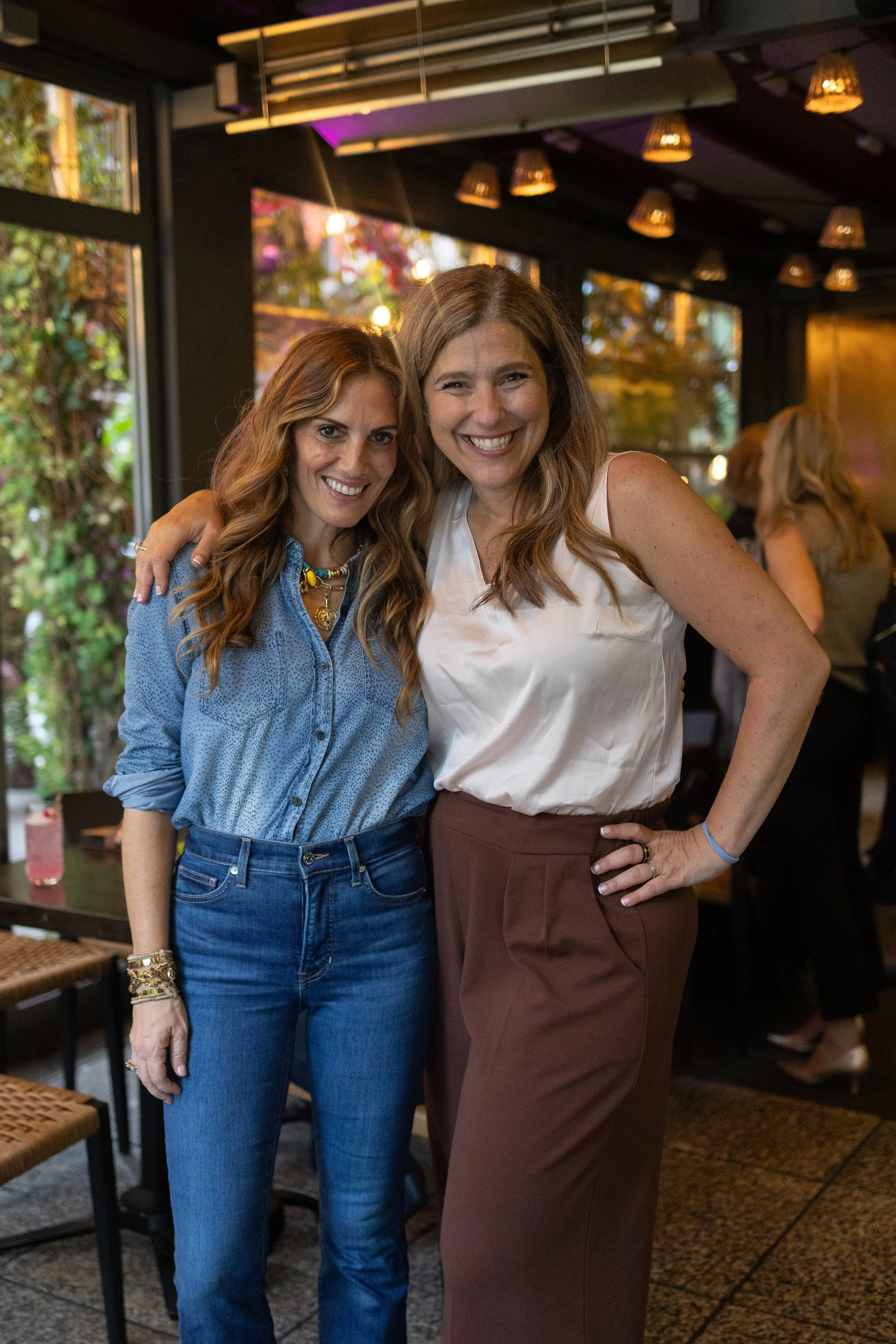 Two women smiling and posing for a photo in a restaurant or cafe. One woman has long, wavy light brown hair and is wearing a blue denim shirt, while the other has long, wavy light brown hair and is wearing a white sleeveless blouse and brown pants. The background shows other people and warm lighting.