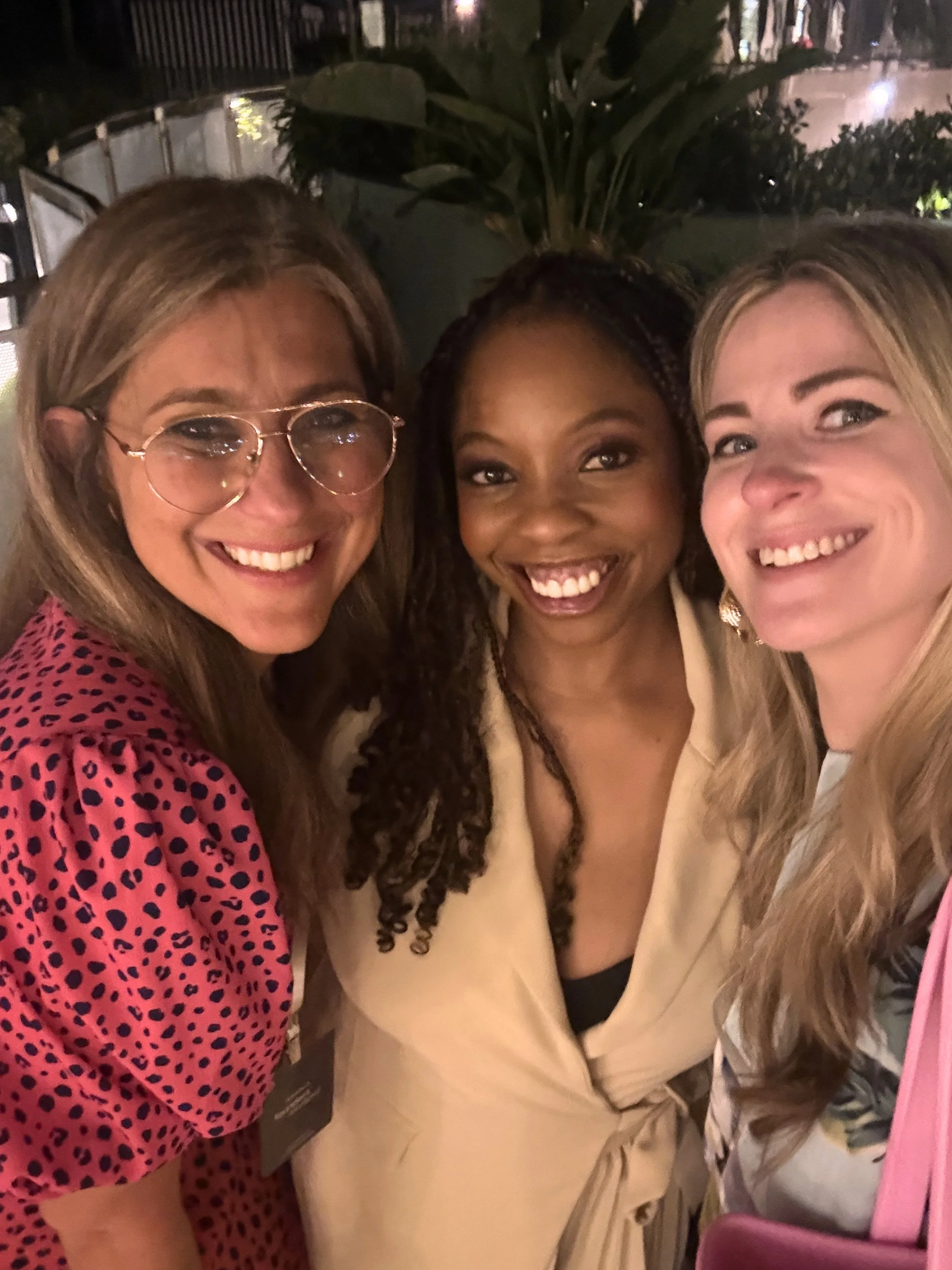 Three women smiling close together in a dimly lit setting, with plants and chairs visible in the background.
