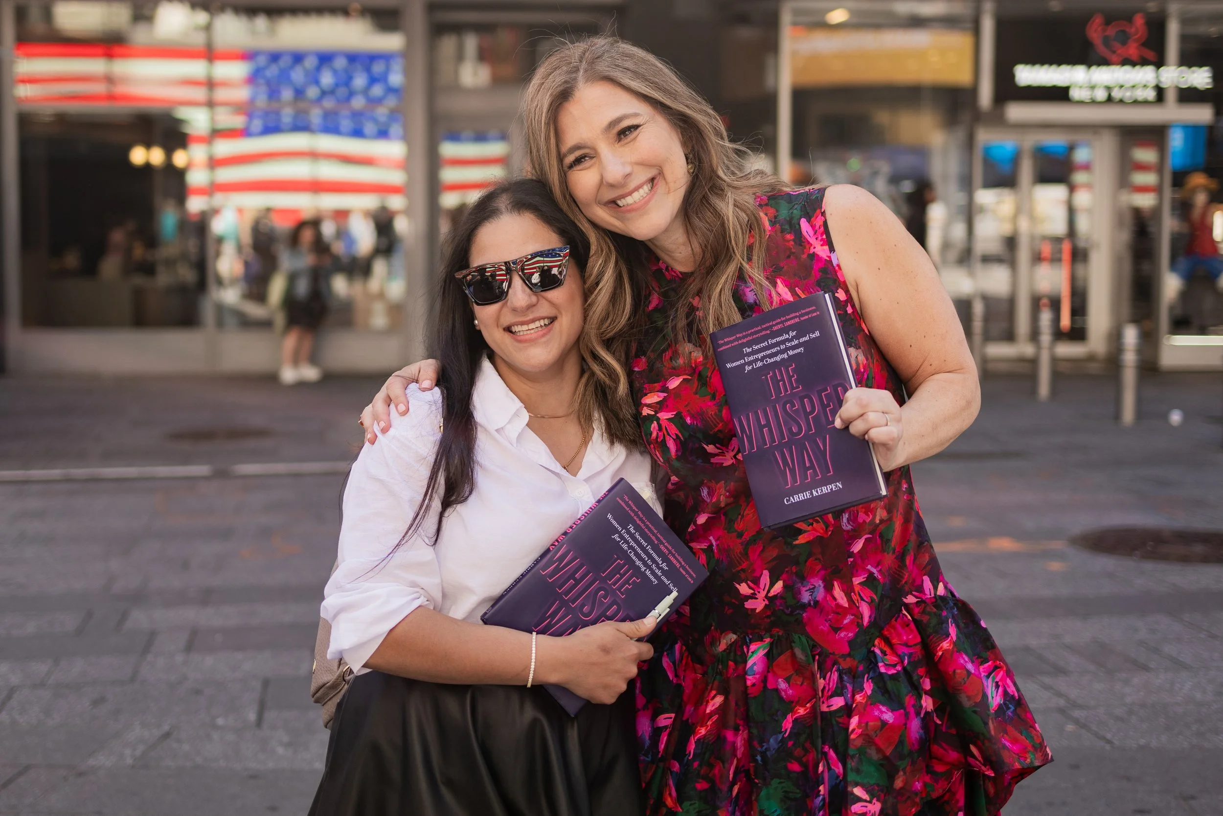 Two women are standing outdoors, smiling, with one woman wearing sunglasses and holding a book. They are embracing each other, and the background features a store with American flags and other people.