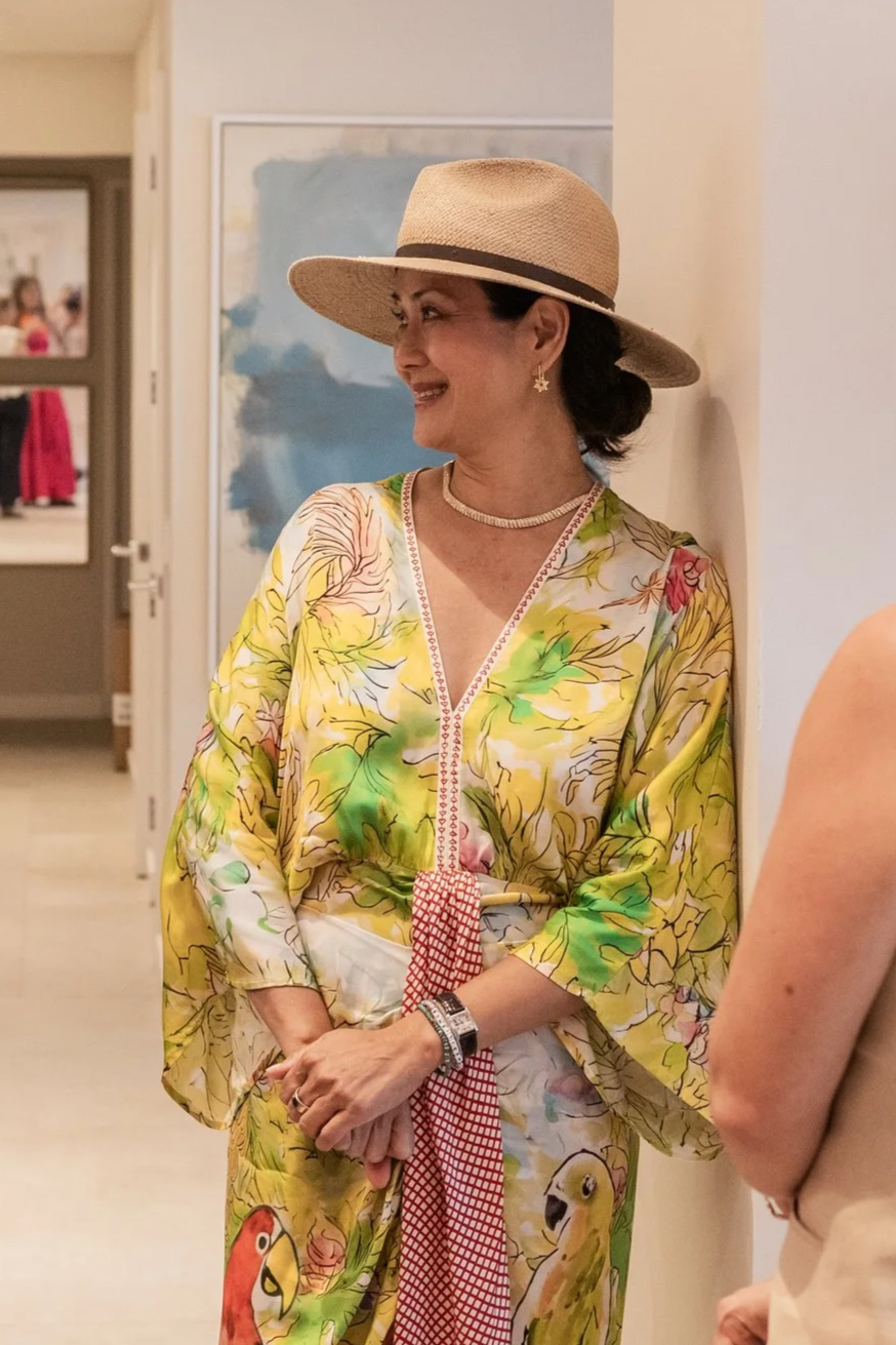 A woman wearing a wide-brimmed straw hat, colorful tropical-patterned kimono, and jewelry is smiling and standing indoors near a wall.