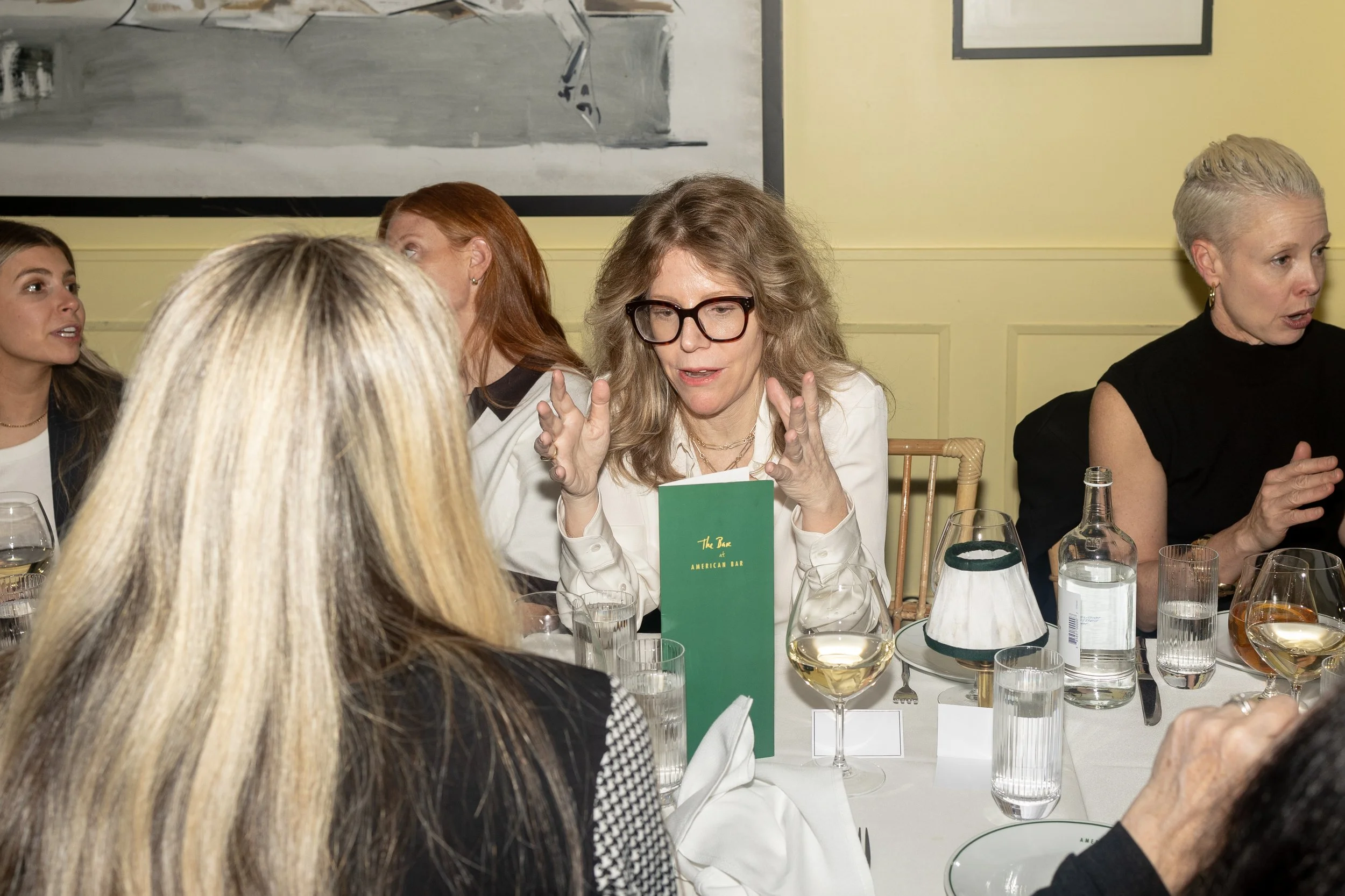 Several women seated at a dinner table having a conversation, with glasses of water and a menu visible on the table, in a restaurant or dining setting.